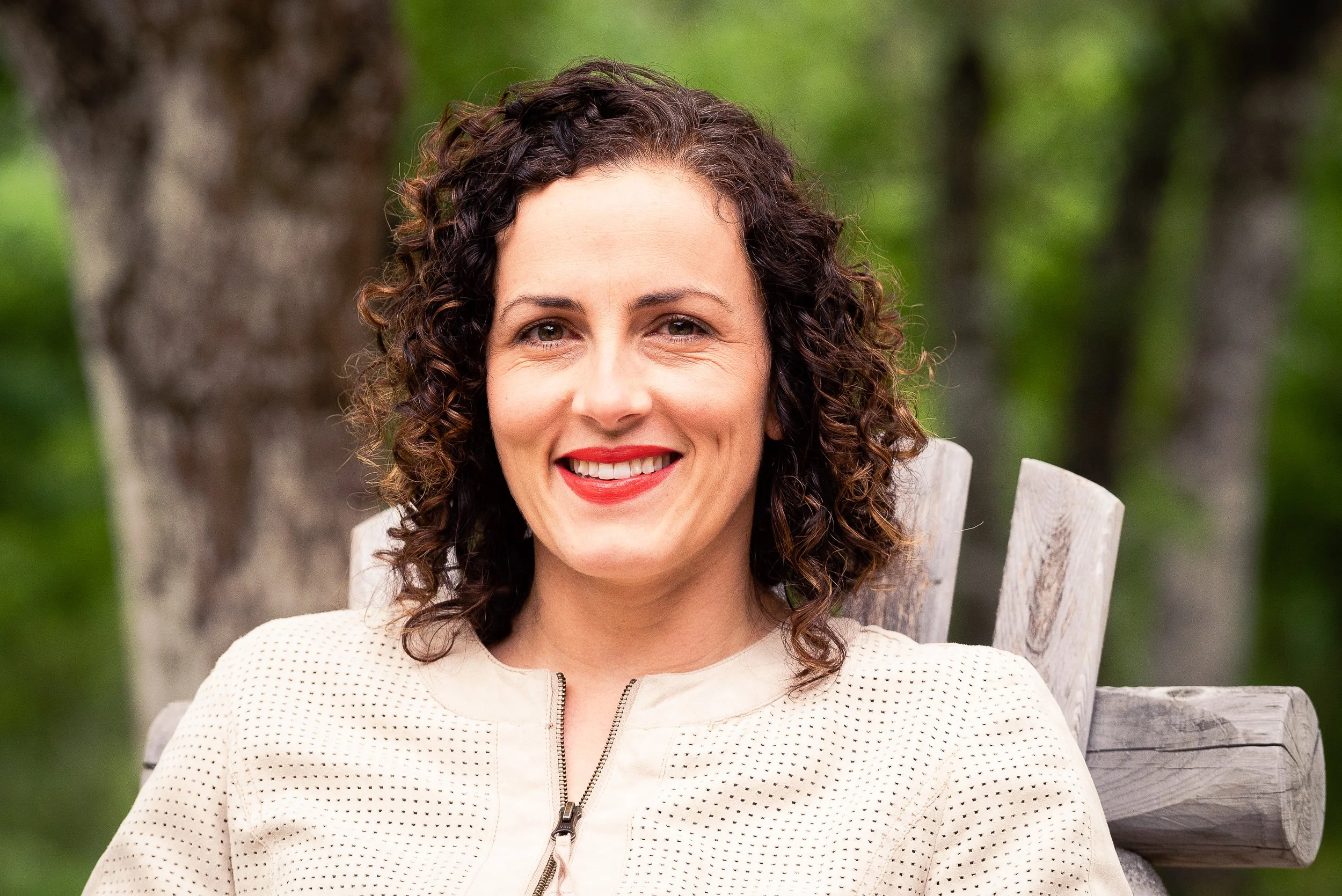 A woman with curly brown hair, wearing red lipstick, smiling, sitting on a wooden chair outdoors with trees and green foliage in the background.