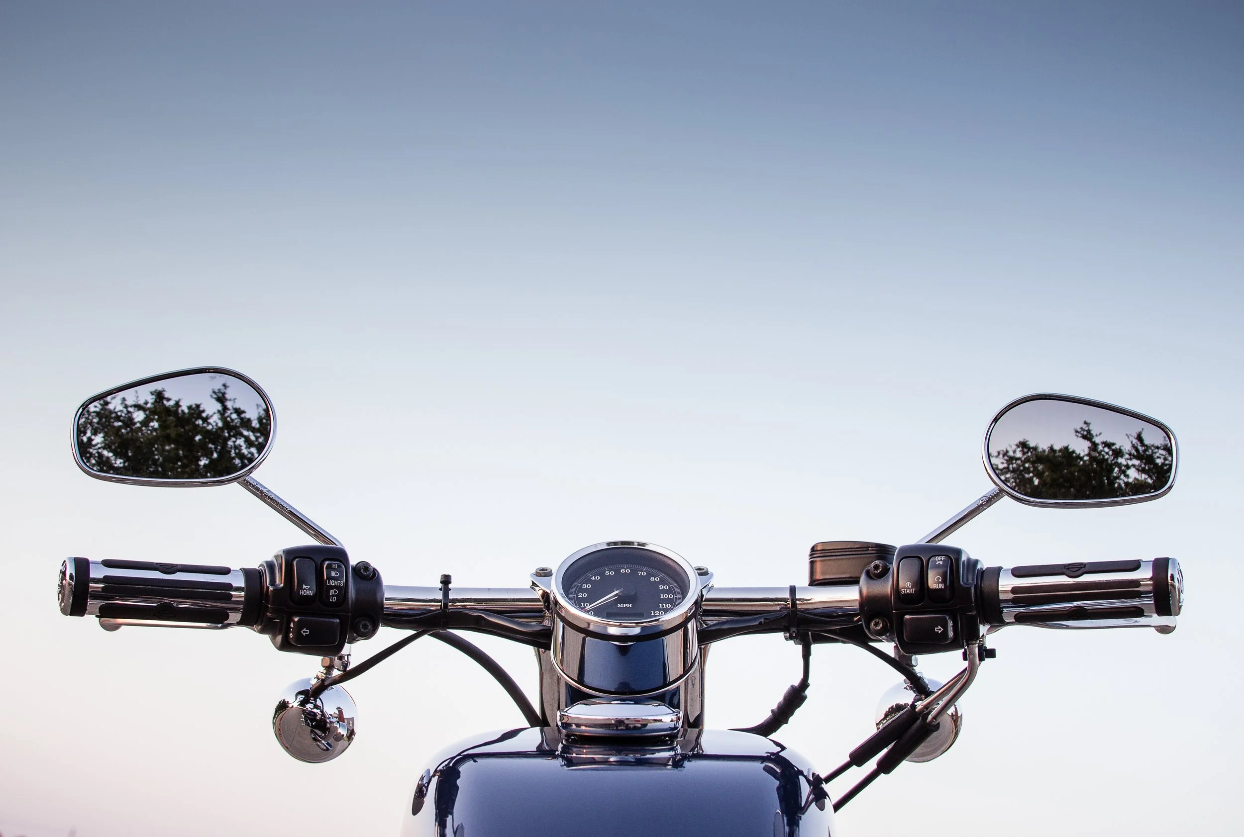 Front view of a motorcycle handlebar with rearview mirrors, speedometer, and controls against a pale blue sky.