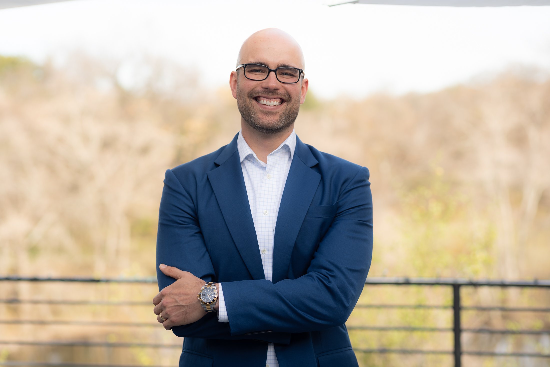 A man outdoors wearing glasses, a blue blazer, and a white shirt, smiling with arms crossed. Background shows trees and a black railing.