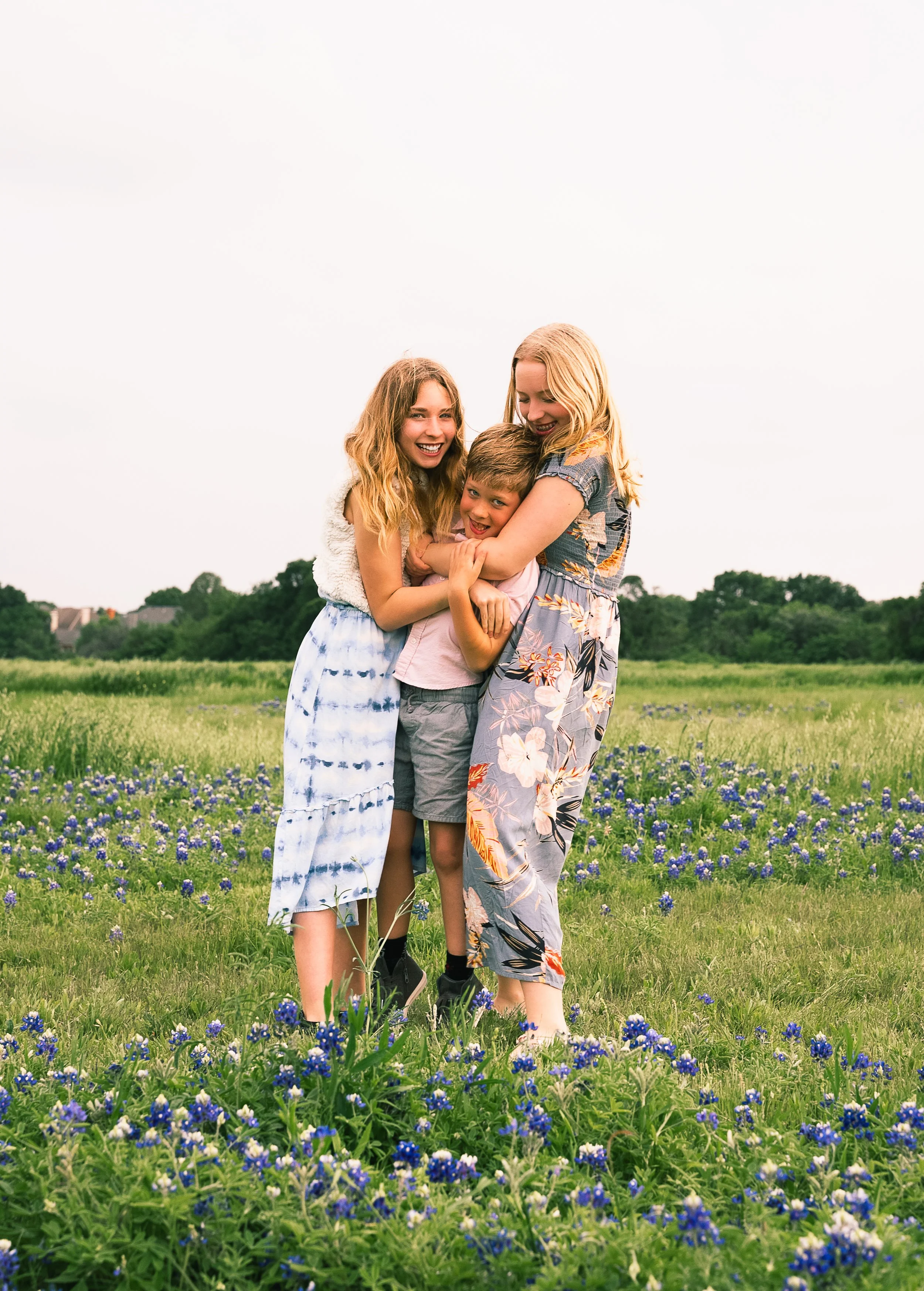 A group of three children, two girls and a boy, embracing each other and smiling in a field of blue flowers with green trees and houses in the background on a cloudy day.