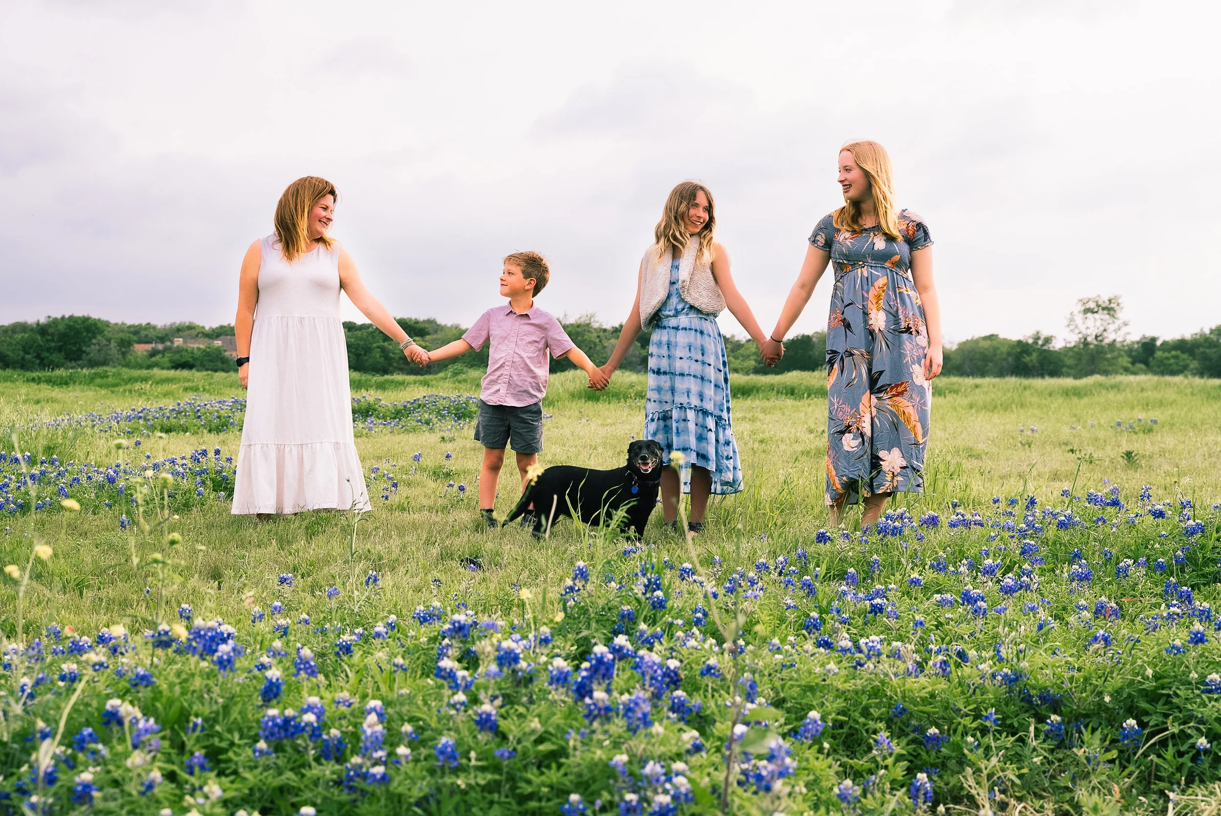 Four women and a boy holding hands in a green field with blue and purple flowers, with a black dog at their feet, under a cloudy sky.