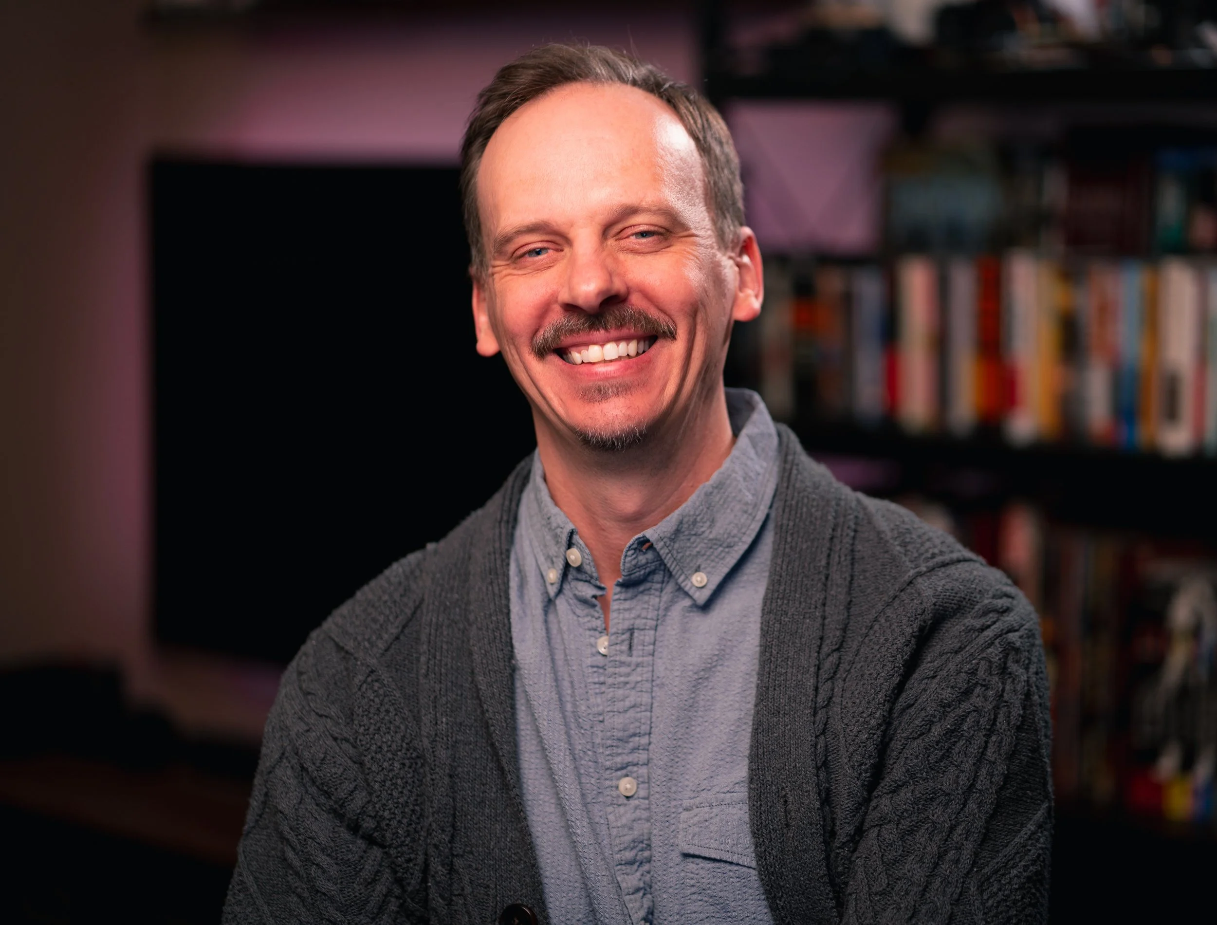 A smiling man with light skin, short brown hair, a mustache, and goatee, wearing a blue shirt and gray cardigan, standing in a room with a bookshelf in the background.