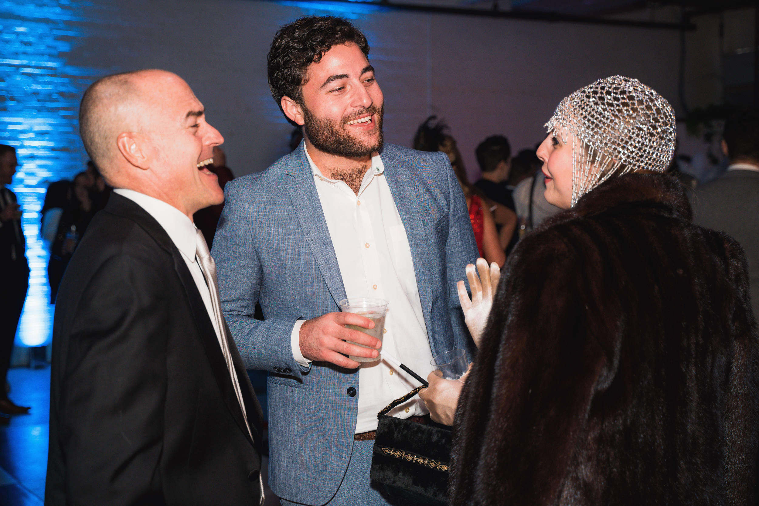 Three people at a social event engaged in conversation. The person on the left is an older man with a bald head and a tuxedo, laughing. The person in the middle is a young man with dark hair and a beard, wearing a blue blazer and white shirt, holding