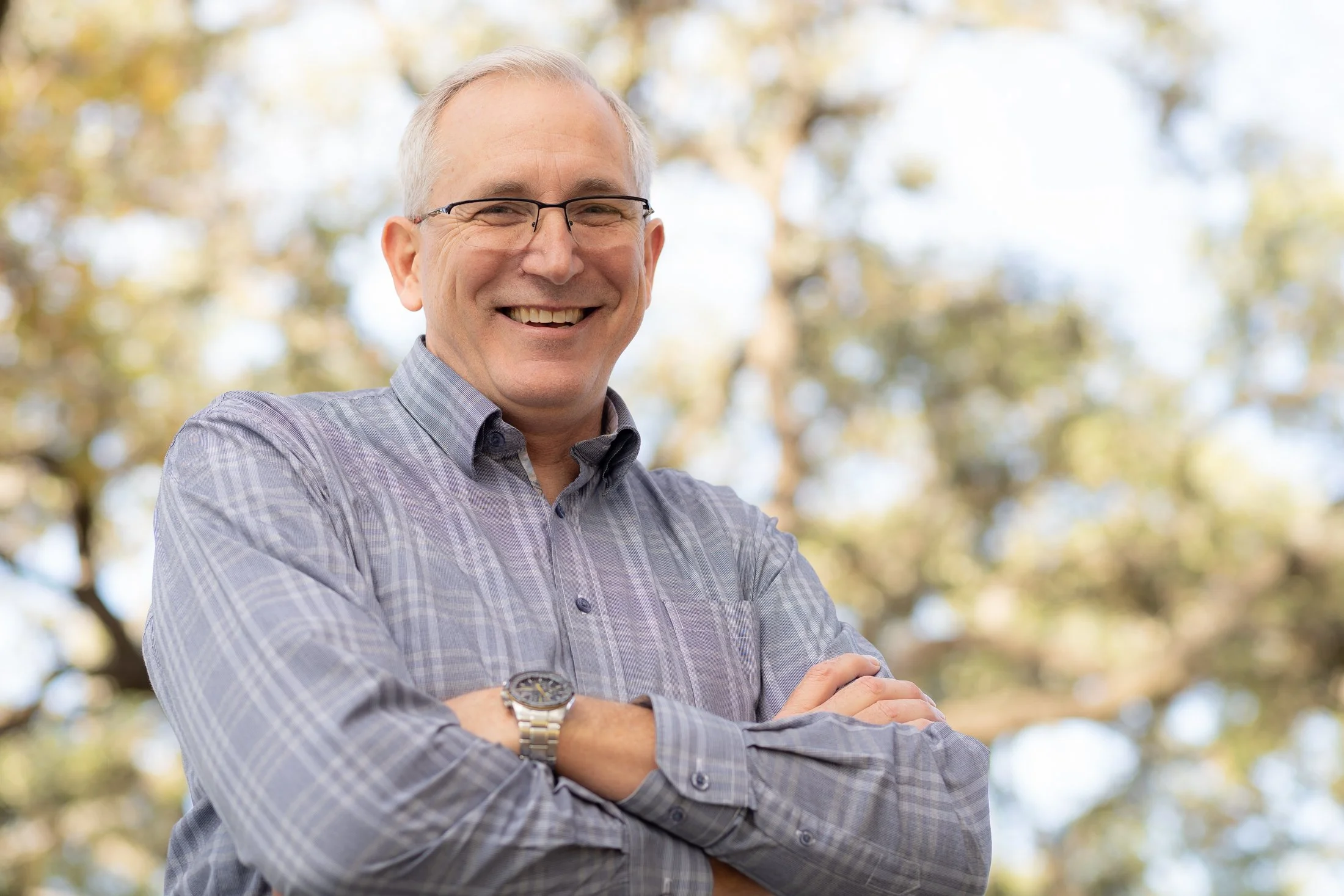 A smiling middle-aged man with gray hair, wearing glasses and a gray checkered shirt, standing outside with trees and blurred leaves in the background.