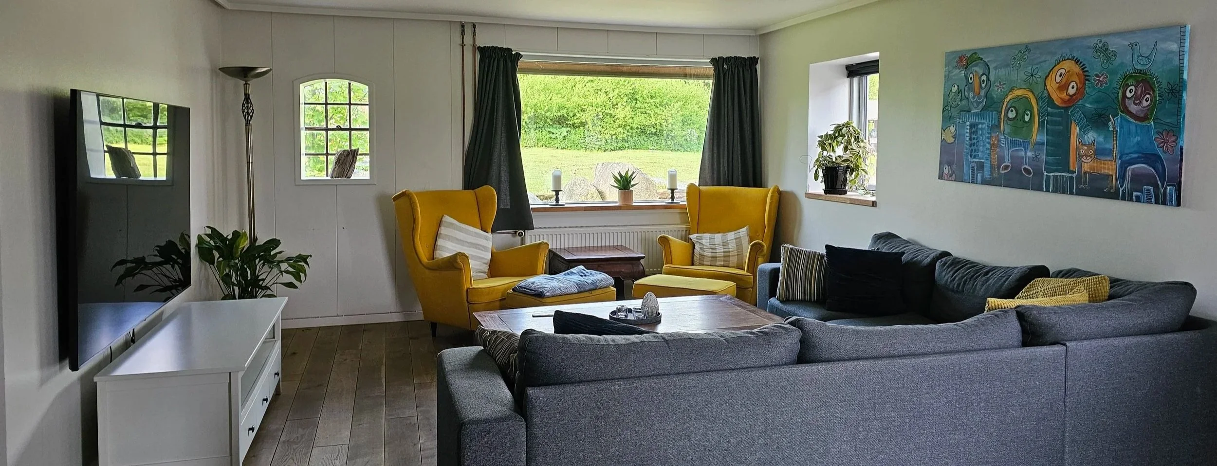 Stue med to store, gule lænestole, en grå sofa med stribede puder, et træbord, et vægkunst med farverige figurer, en hvid medielįnk, grønne planter, vinduer med grønne gardiner, udsigt til grønne marker udenfor.