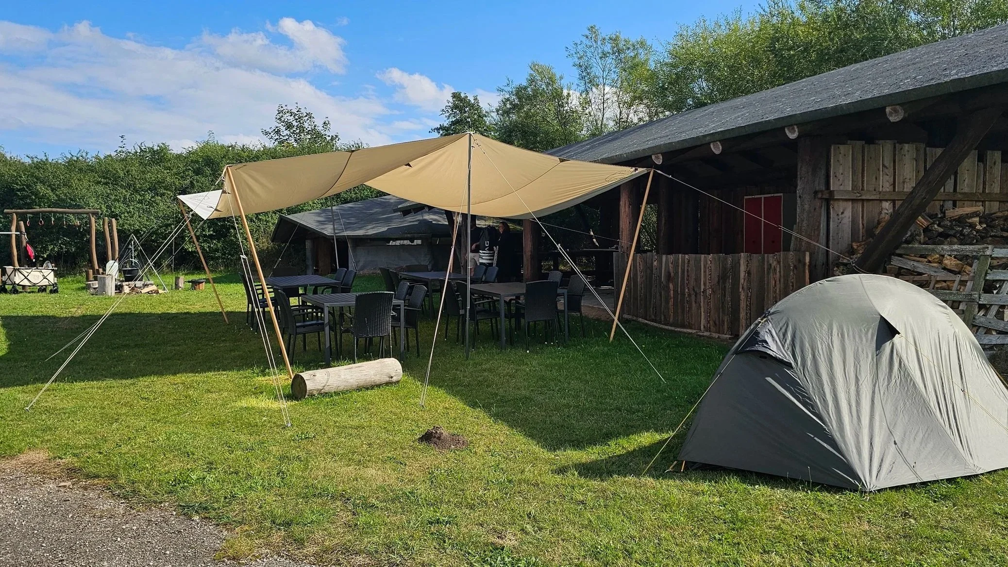 En campingplads med telt, borde, stole og en tarps, omgivet af grønne træer og bygninger.