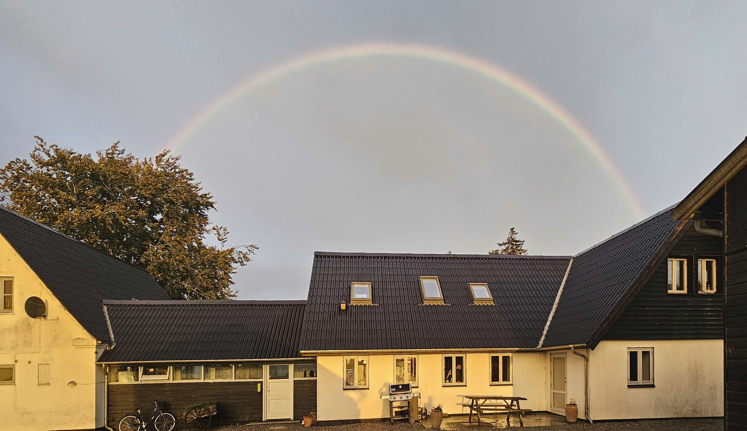 Hus med sort tag og skur med hvid væg og udendørsbord foran, med regnbue i klar himmel