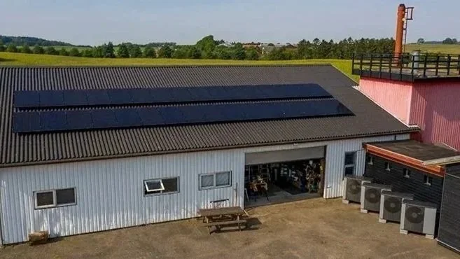 En stor industriel bygning med solceller på taget, udendørs område med et picnicbord og flere luftkonditioneringsenheder, omgivet af grønne marker.
