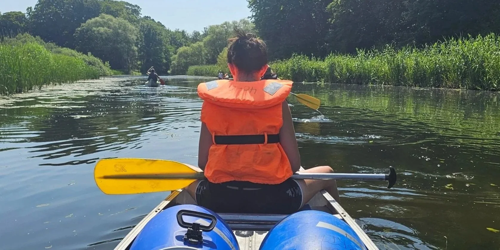 Person i orange redningsvest padler i kano på en rolig flod omgivet af grønt vegetation og træer på en solrig dag.