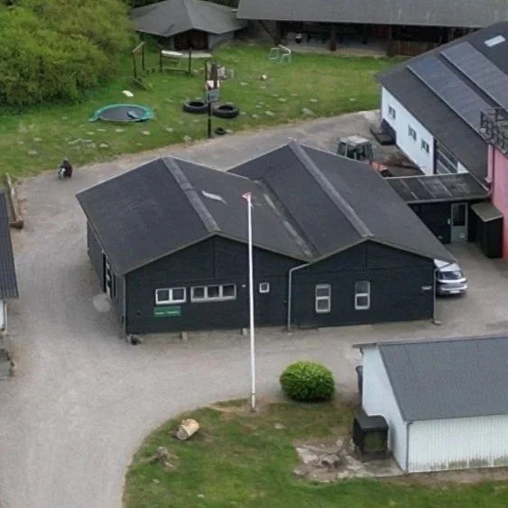 Et sort træhus med flere sektioner set fra luften, omgivet af græs, en sti og omkringliggende bygninger, en bil er parkeret ved huset.