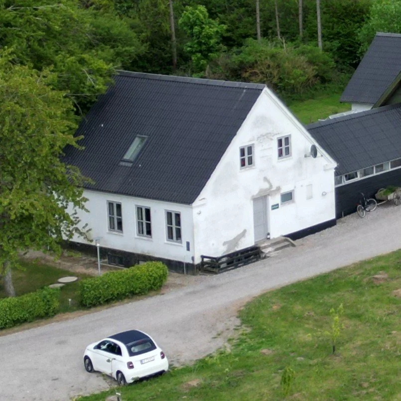 Landsbyhus med sort tag, hvid facade, omgivet af grønne træer og græs, en parkeret hvid bil foran, cykel ved husets side.