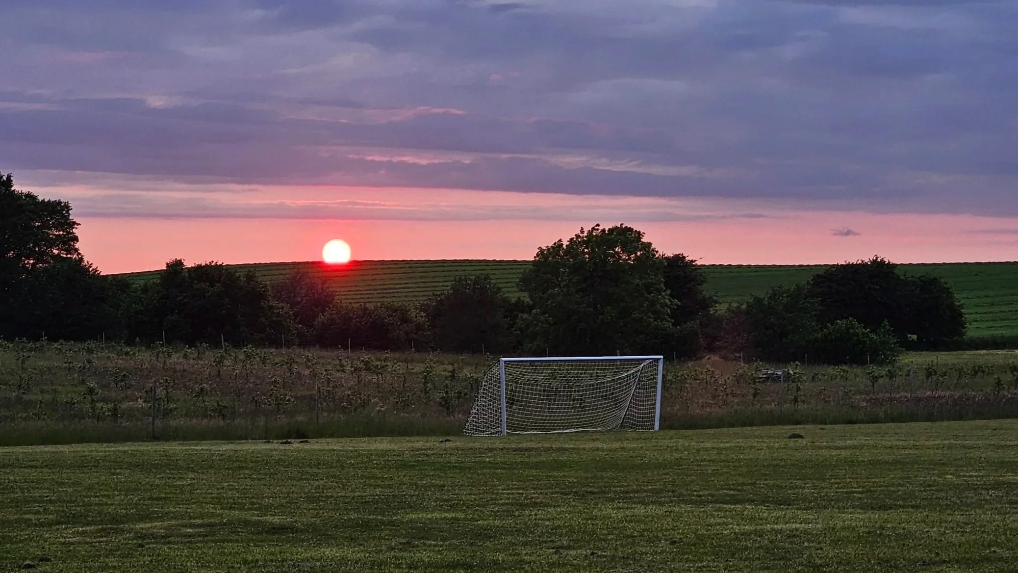 Solnedgang over et grønt fodboldfelt med mål, omgivet af marker og træer, med en farverig himmel i baggrunden.
