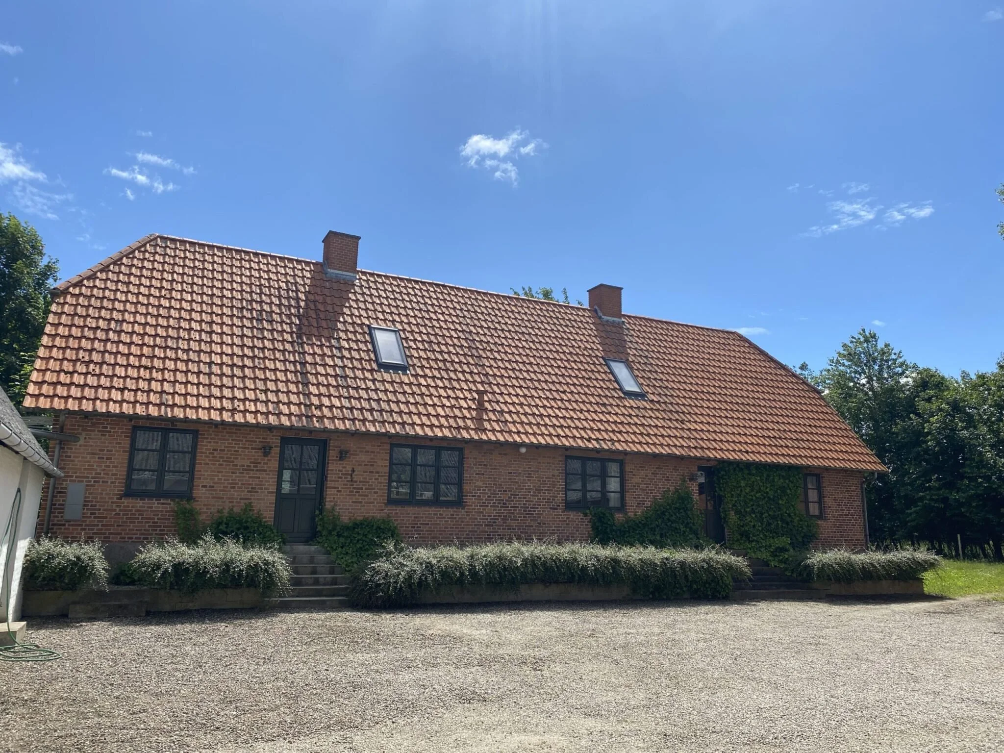 En lang rødbrun murstensbygning med tag af røde teglsten, to skorstene, to skydedøre med vinduer, og små buske foran indgangen. Omkring bygningen er der træer og en græsplæne, under en klar blå himmel med få spredte skyer.