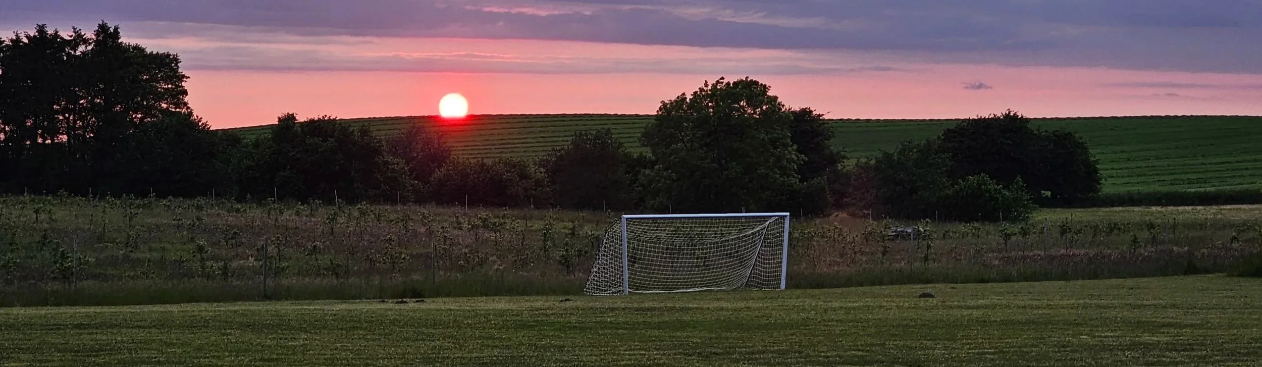 Solnedgang over et grønt fodboldmål på en græsplet, med marker og træer i baggrunden.