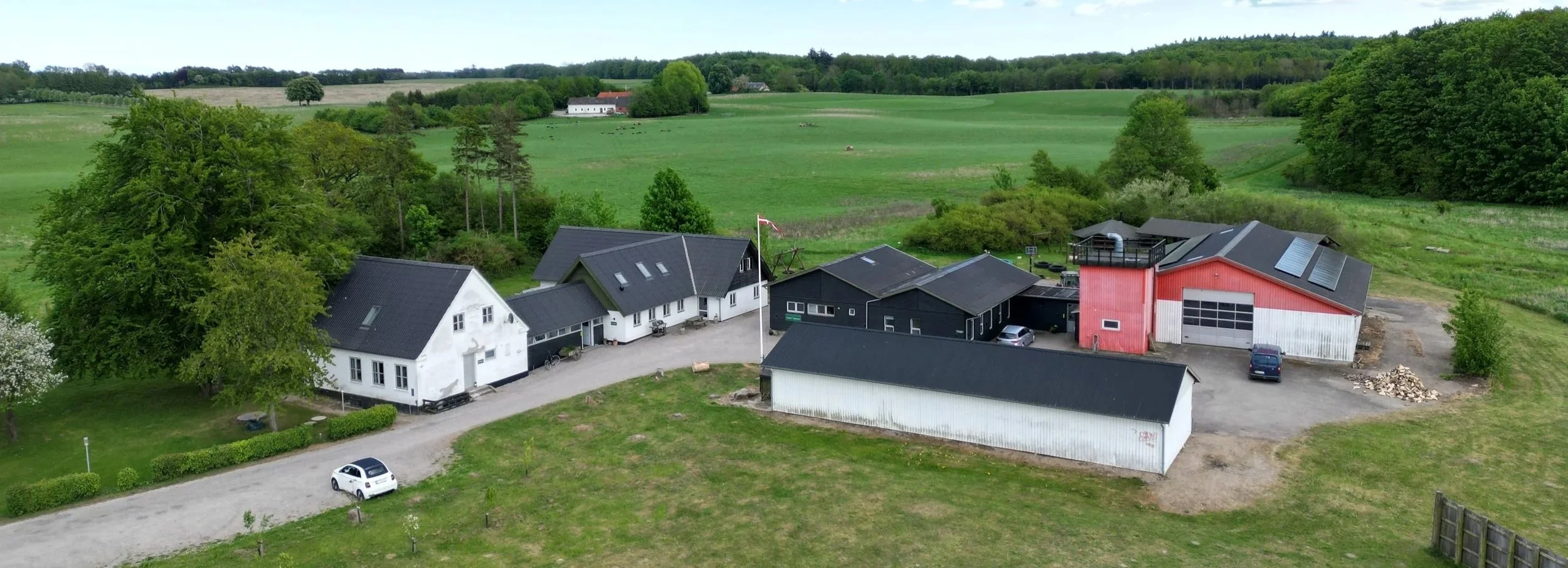 Luftfoto af en gård med flere huse, tre større bygninger, biler og traktor, omgivet af grønne marker og træer.