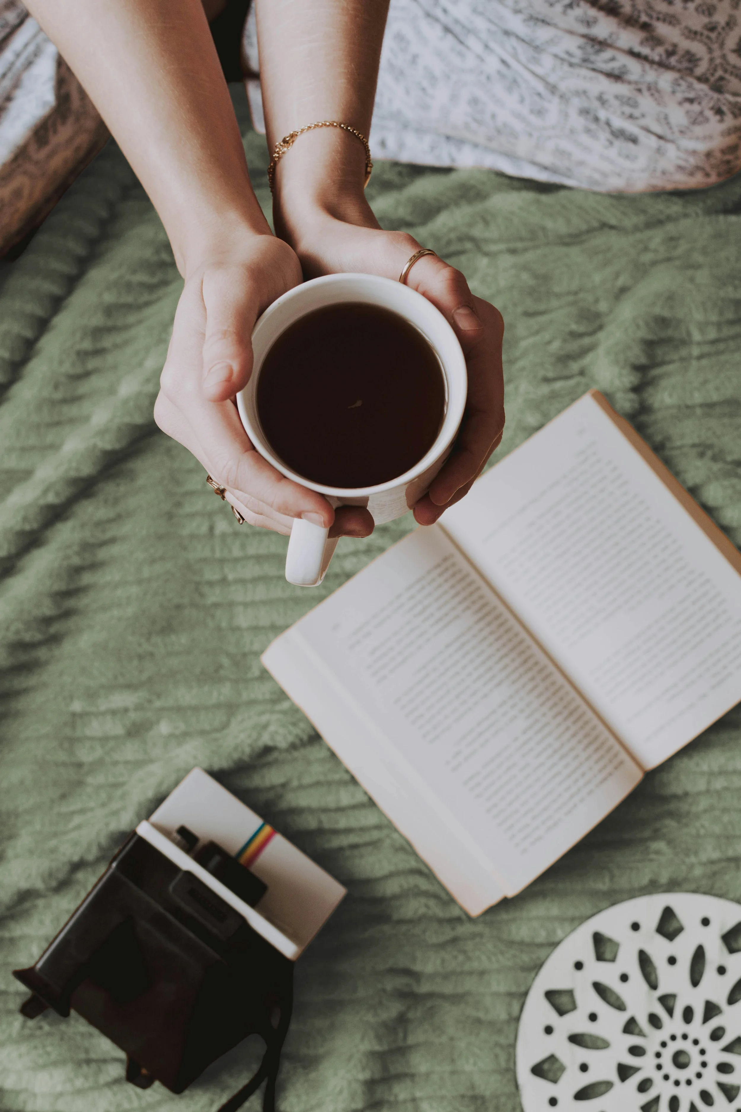 Person holding a mug of coffee above an open book, with a camera and decorative plate on a green textured blanket.
