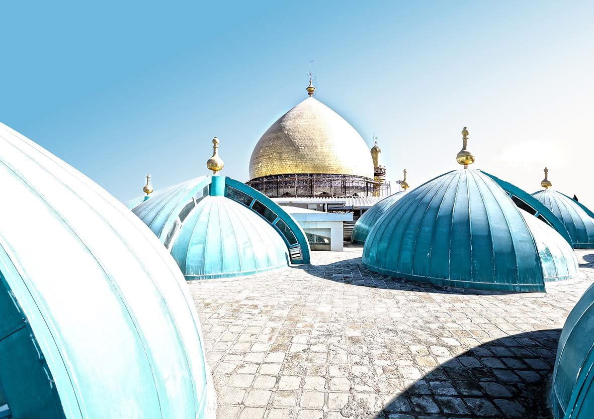 View of the Shrine of Imam Ali (as) with gold and blue domes against a clear blue sky.