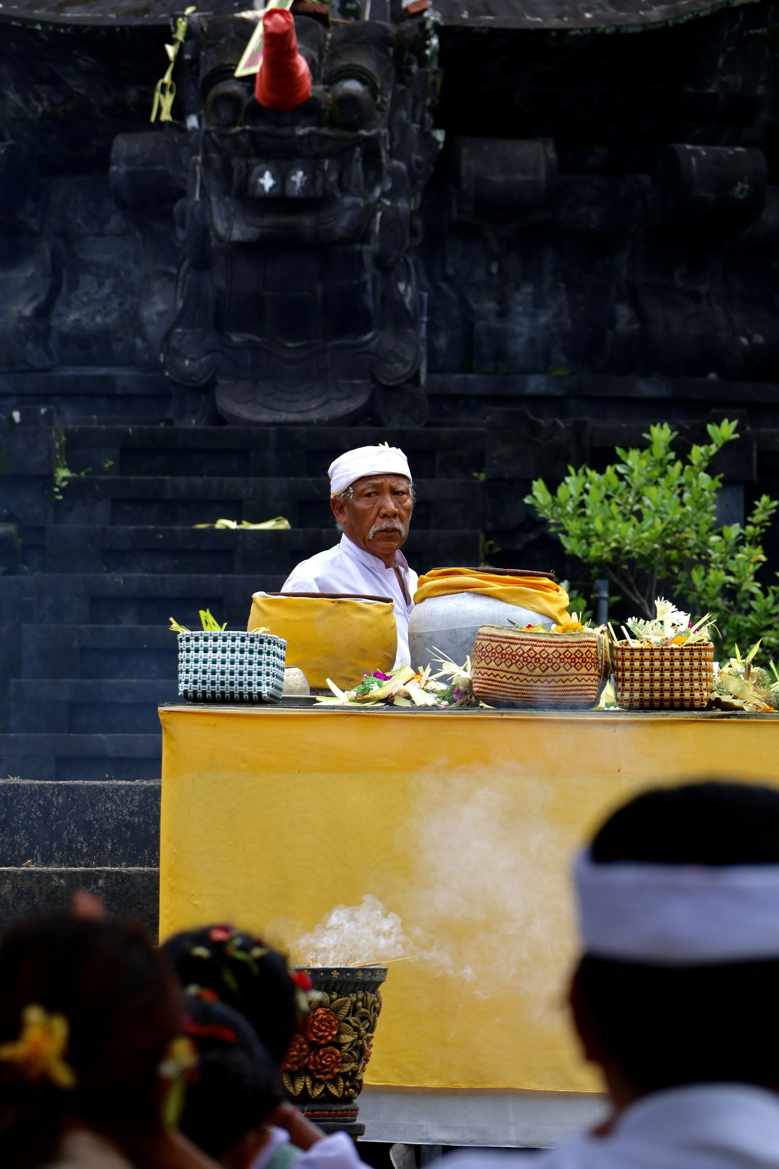 An elderly man dressed in white, participating in a traditional ceremony at a temple with cultural artifacts and offerings on a yellow cloth table, background features dark stone carvings.