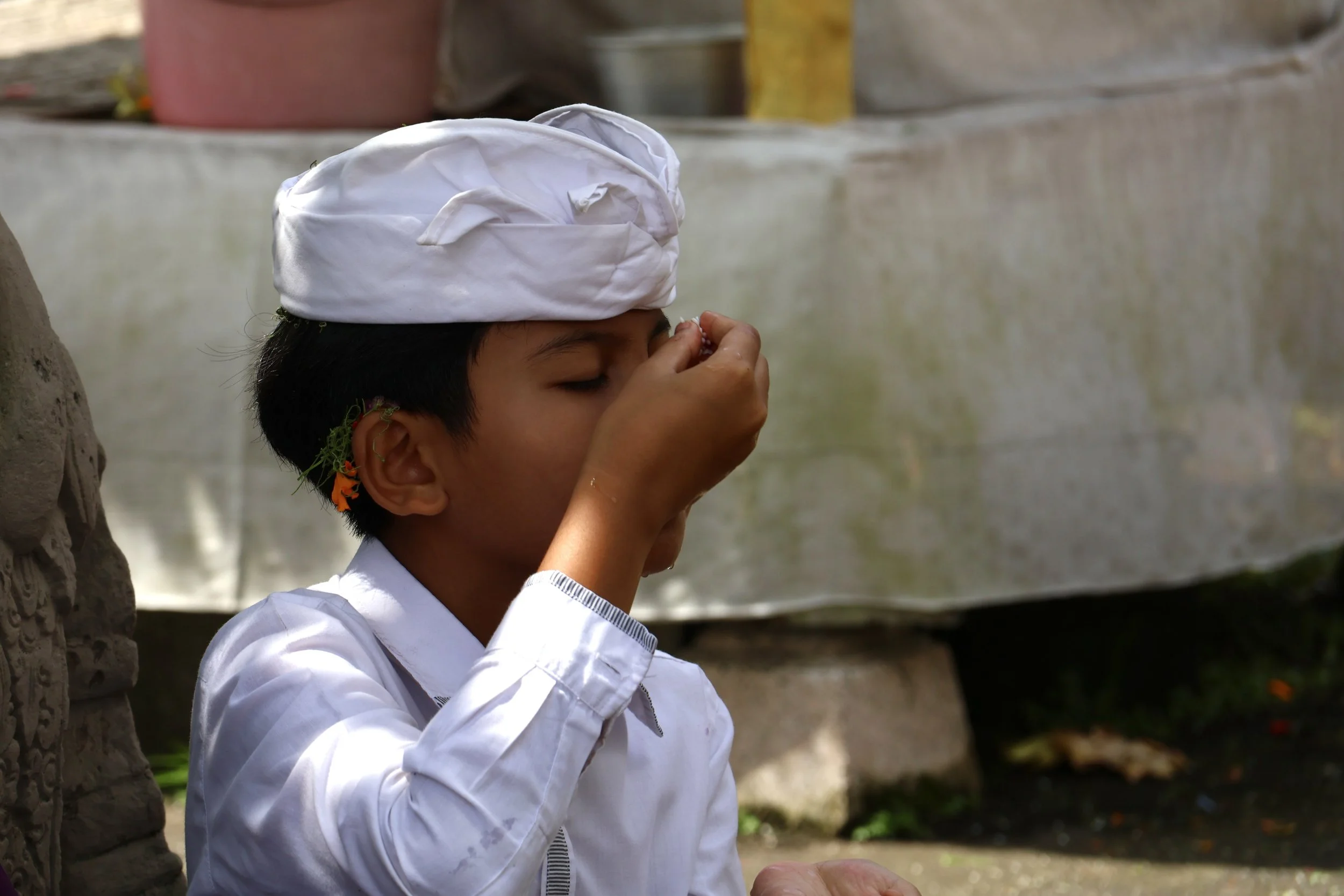 Young Balinese boy dressed in a white shirt and a traditional white cap is closing his eyes and touching his face, standing outdoors near a stone structure with blurred background.