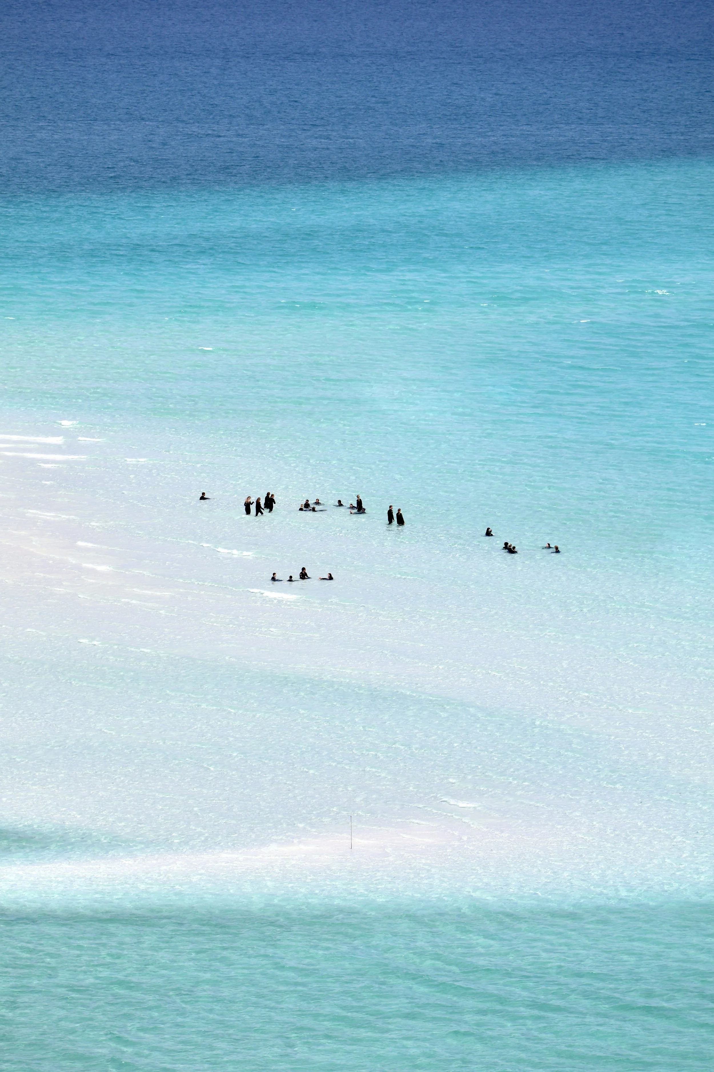 People in black wetsuits swimming and floating in a turquoise ocean with a white sandy beach.