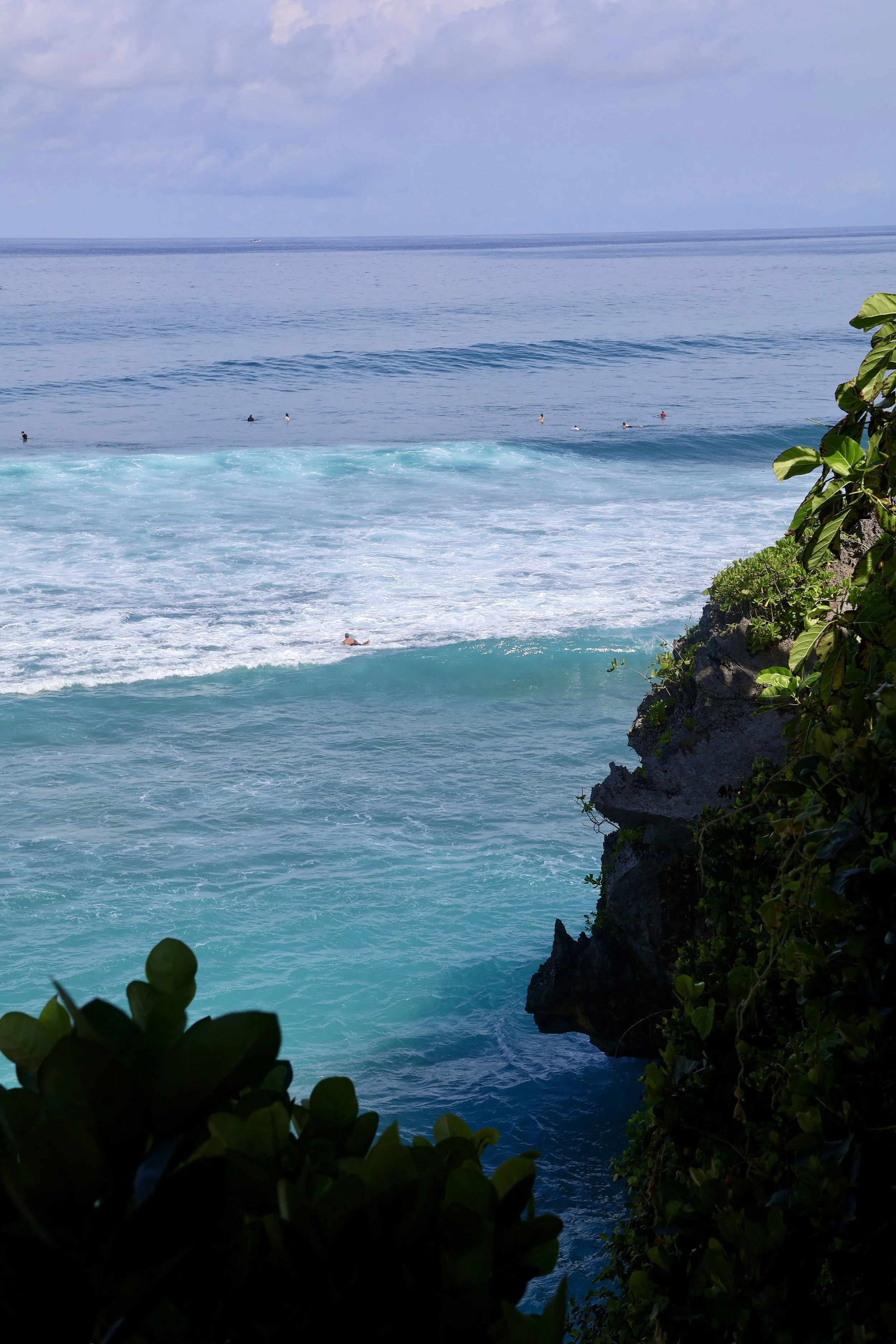 Ocean view from a rocky cliff with green foliage in the foreground, showing waves and surfers in the water and a cloudy sky above.