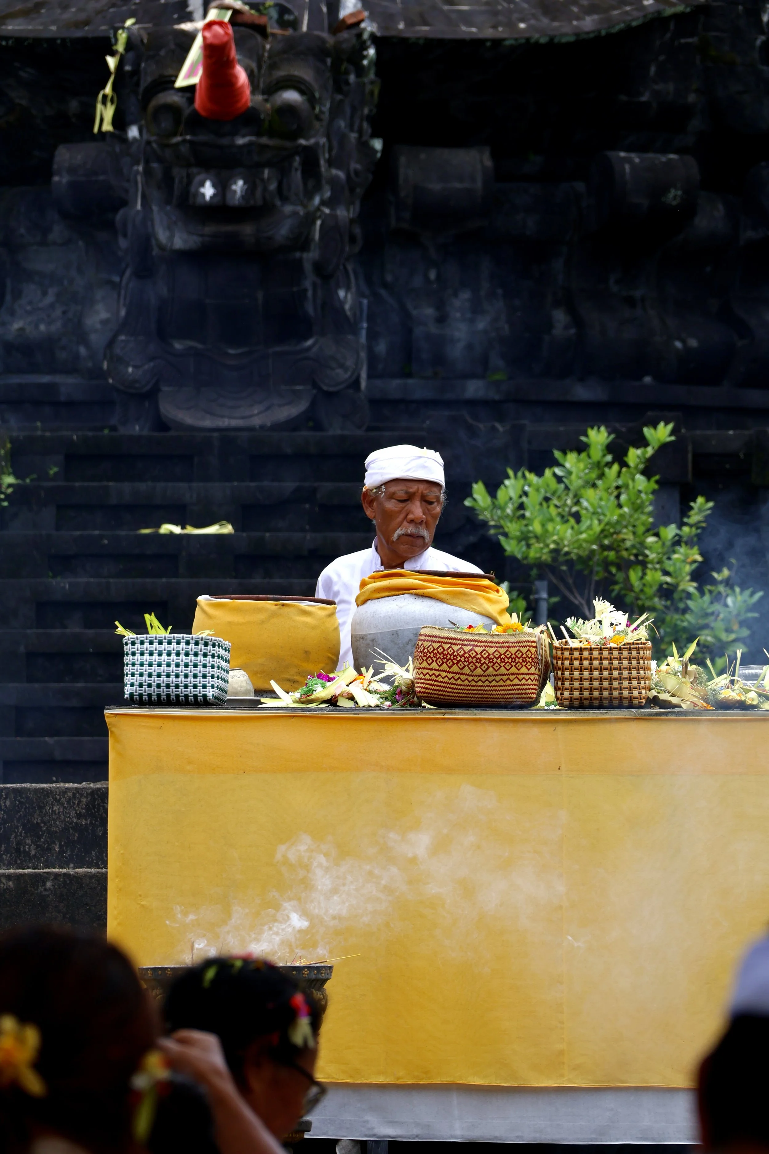 A Balinese man dressed in traditional white attire and a white headscarf is performing a ceremonial ritual at a yellow altar, surrounded by various offerings and flowers, with a dark stone sculpture of a mythical creature in the background.