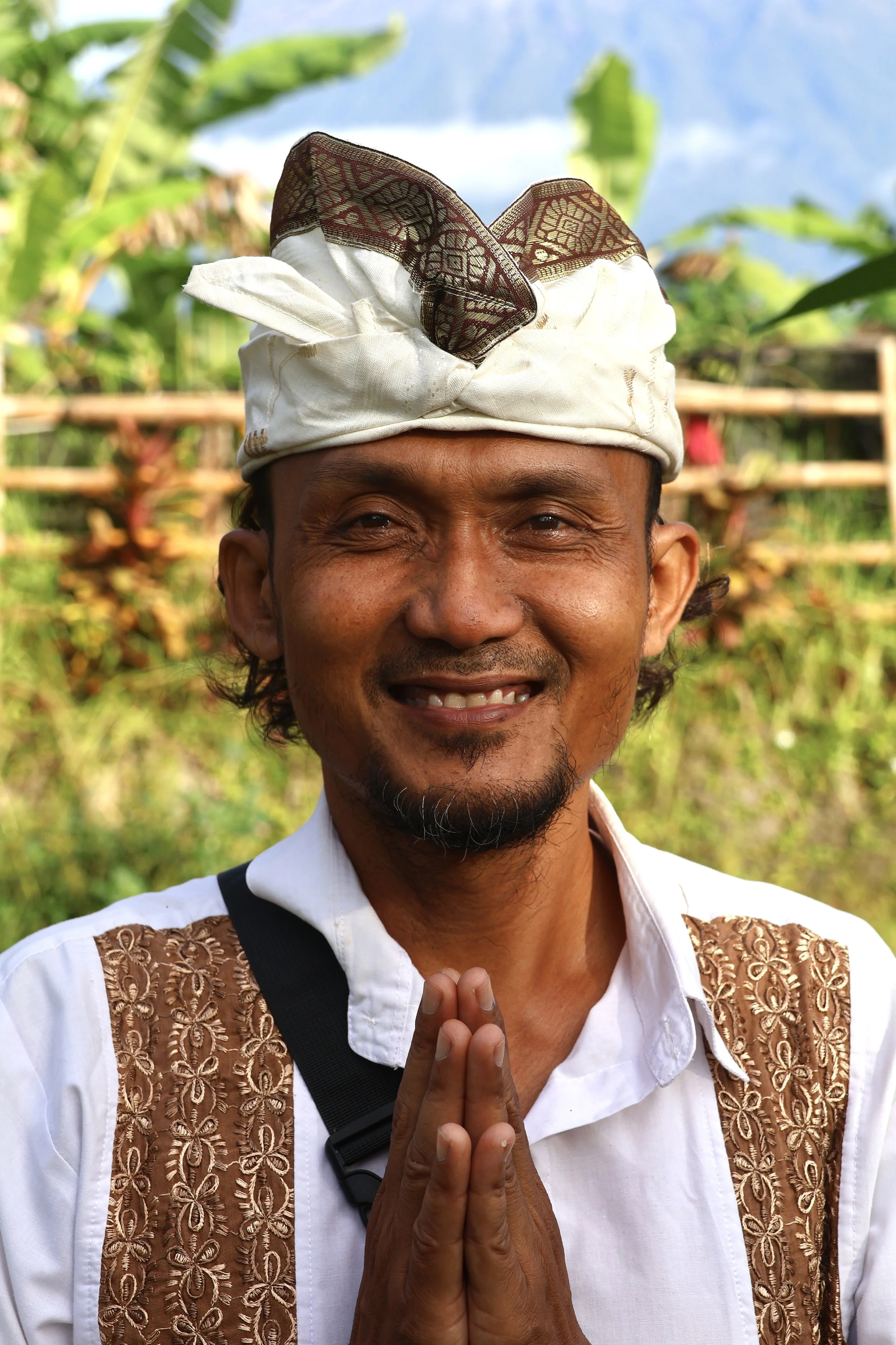 A smiling man wearing traditional Balinese attire, including a white shirt with embroidered brown accents and a white headwrap, standing outdoors with lush green foliage in the background.