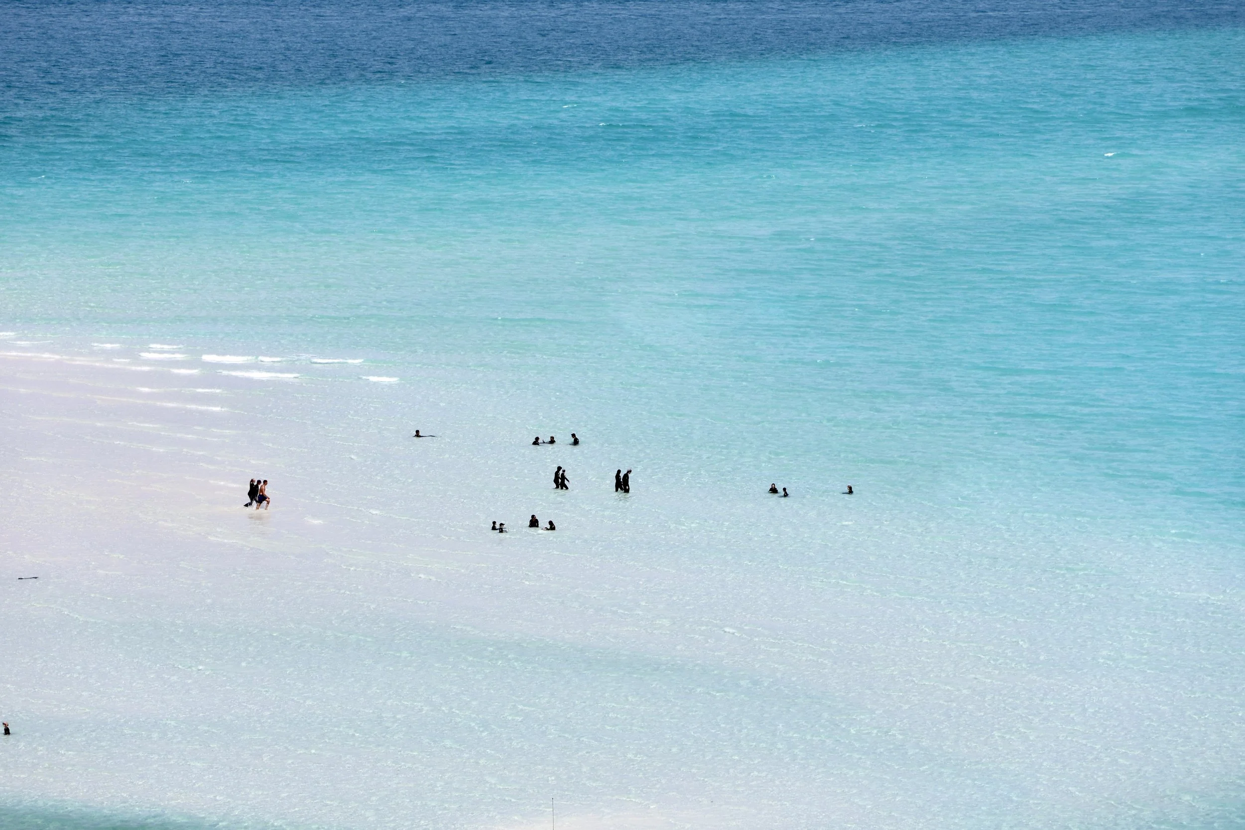 People swimming and walking at the beach, with turquoise water and white sand.