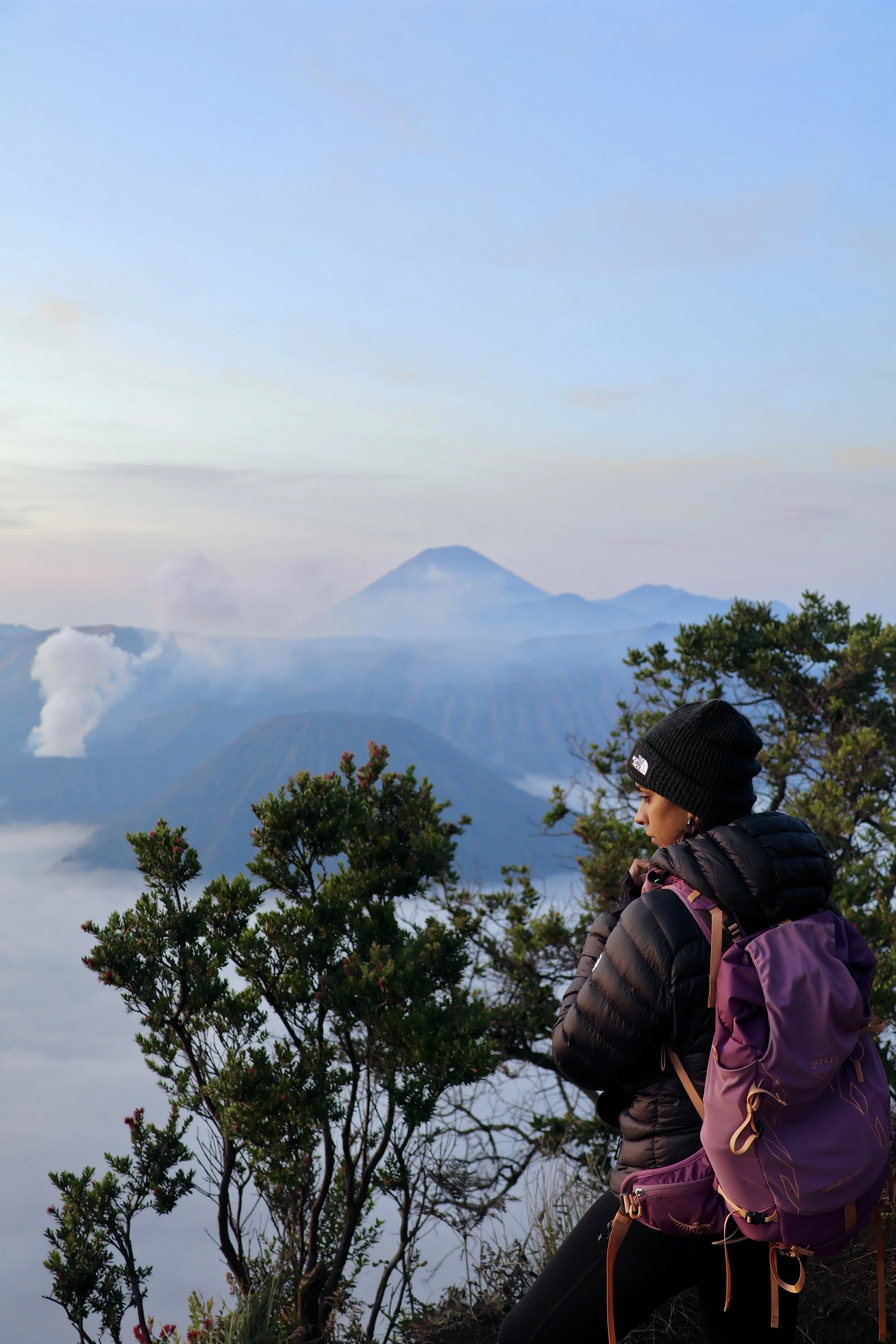 A woman wearing a black jacket, purple backpack, and black beanie standing on a mountain trail, overlooking misty mountains and volcanoes with clouds and a blue sky in the background.