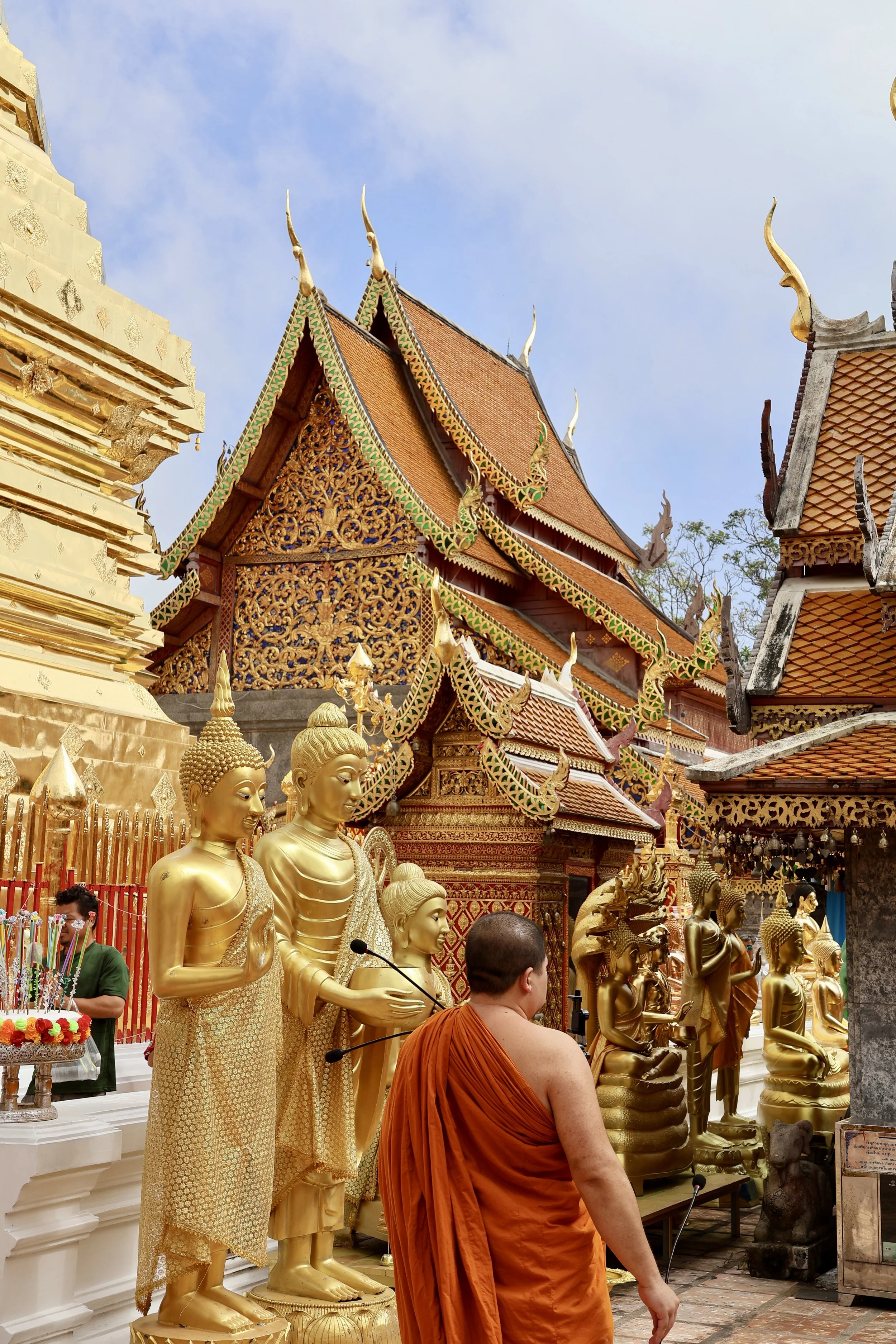 Monk in orange robes observing gold Buddha statues inside a richly decorated temple with traditional Thai architecture.