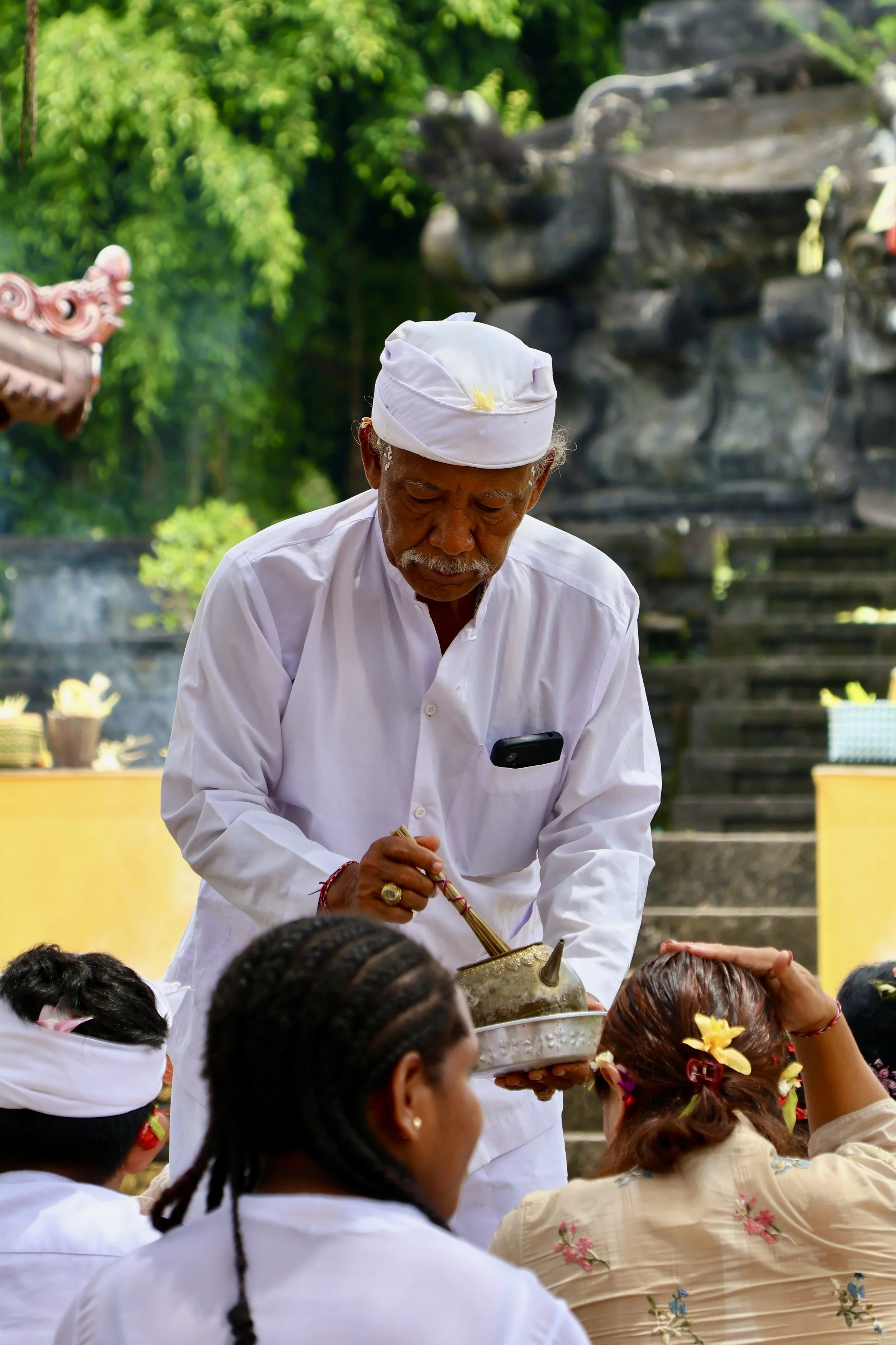 An elderly Balinese man in traditional white attire and headwear conducts a spiritual ritual or blessing with a container and incense in front of a group of women.