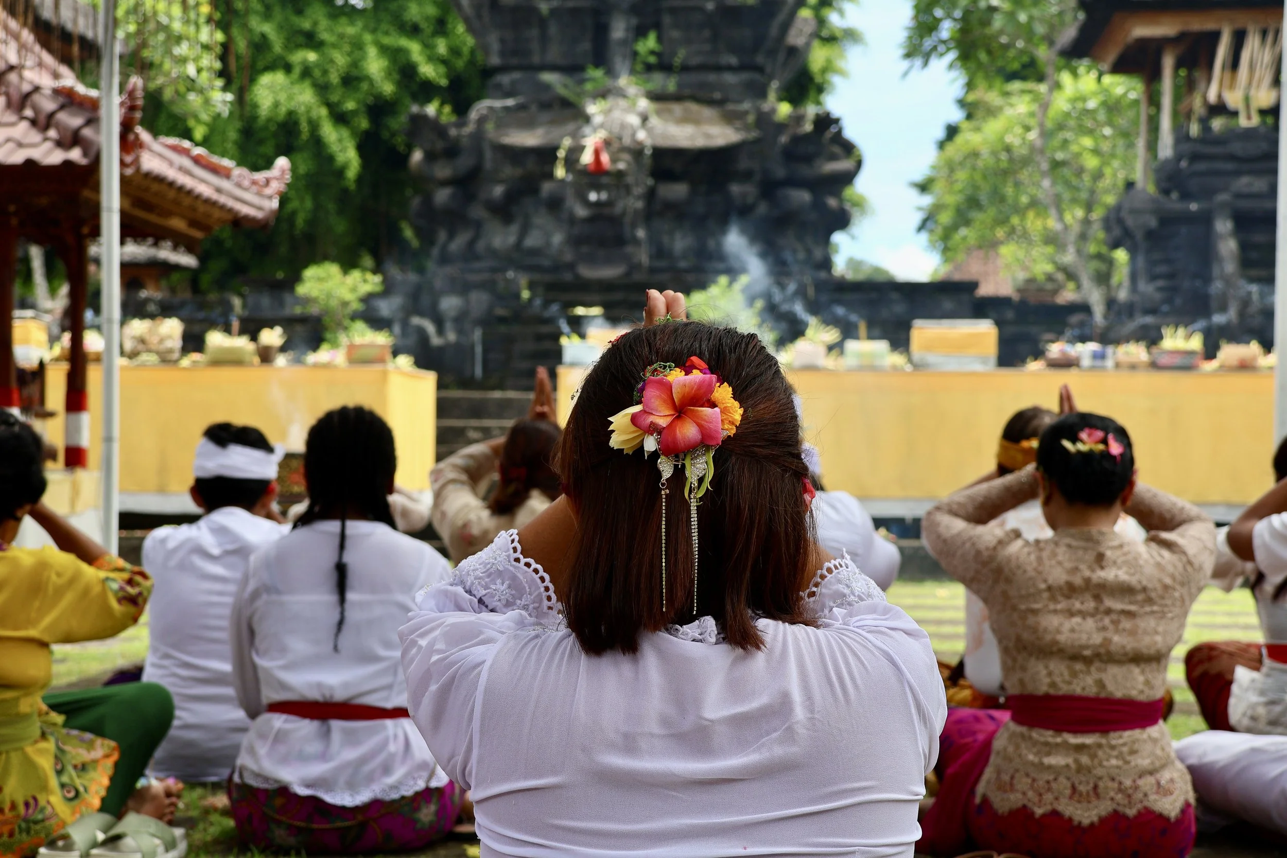 People sitting on the ground during a traditional Balinese ceremony at a temple, with a large black shrine or statue in the background, and lush green trees surrounding the scene.