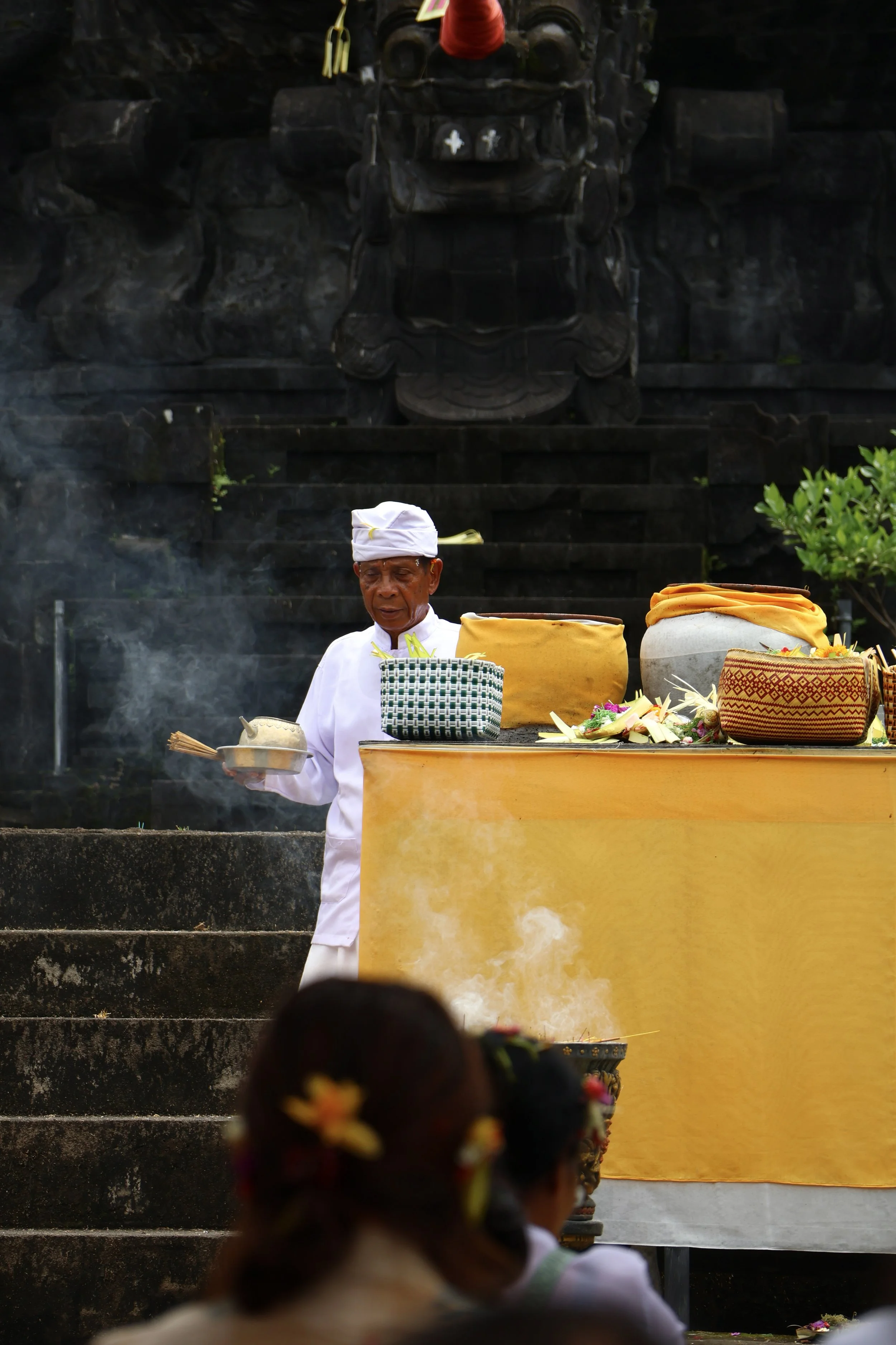 A Balinese priest dressed in white, performing a traditional ceremony at a yellow altar with offerings and incense in front of a dark stone wall with carved details.