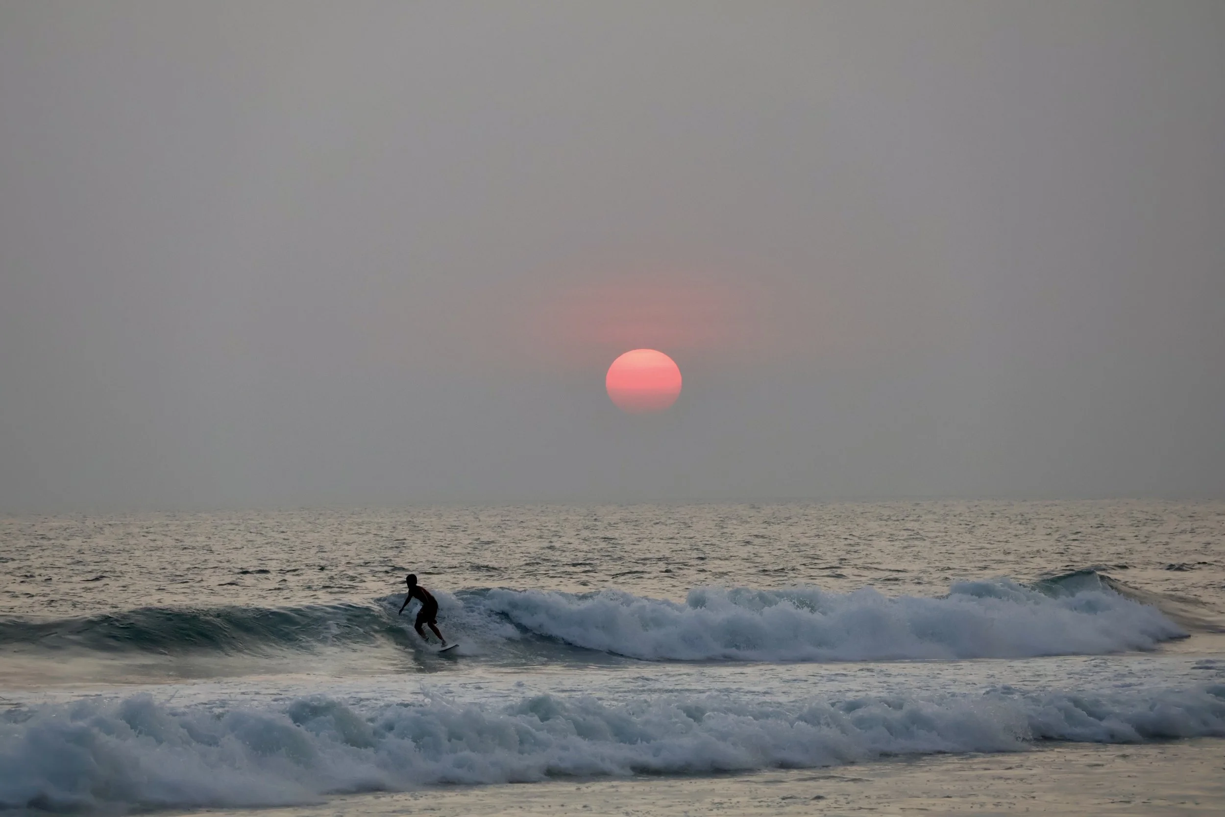 A person surfing on a wave during sunset over the ocean, with a pink and orange sky.