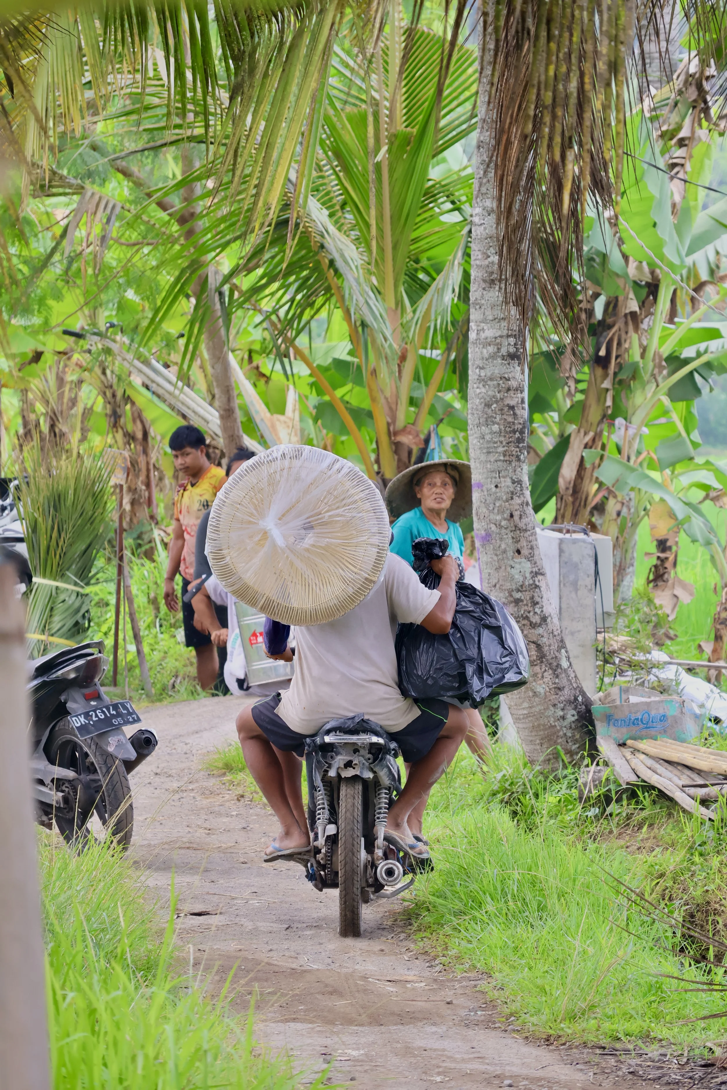 A man riding a motorcycle with a large woven hat on his head, carrying a black plastic bag, on a dirt road surrounded by lush tropical plants, with a woman wearing a wide-brimmed hat standing nearby, and several people walking in the background.