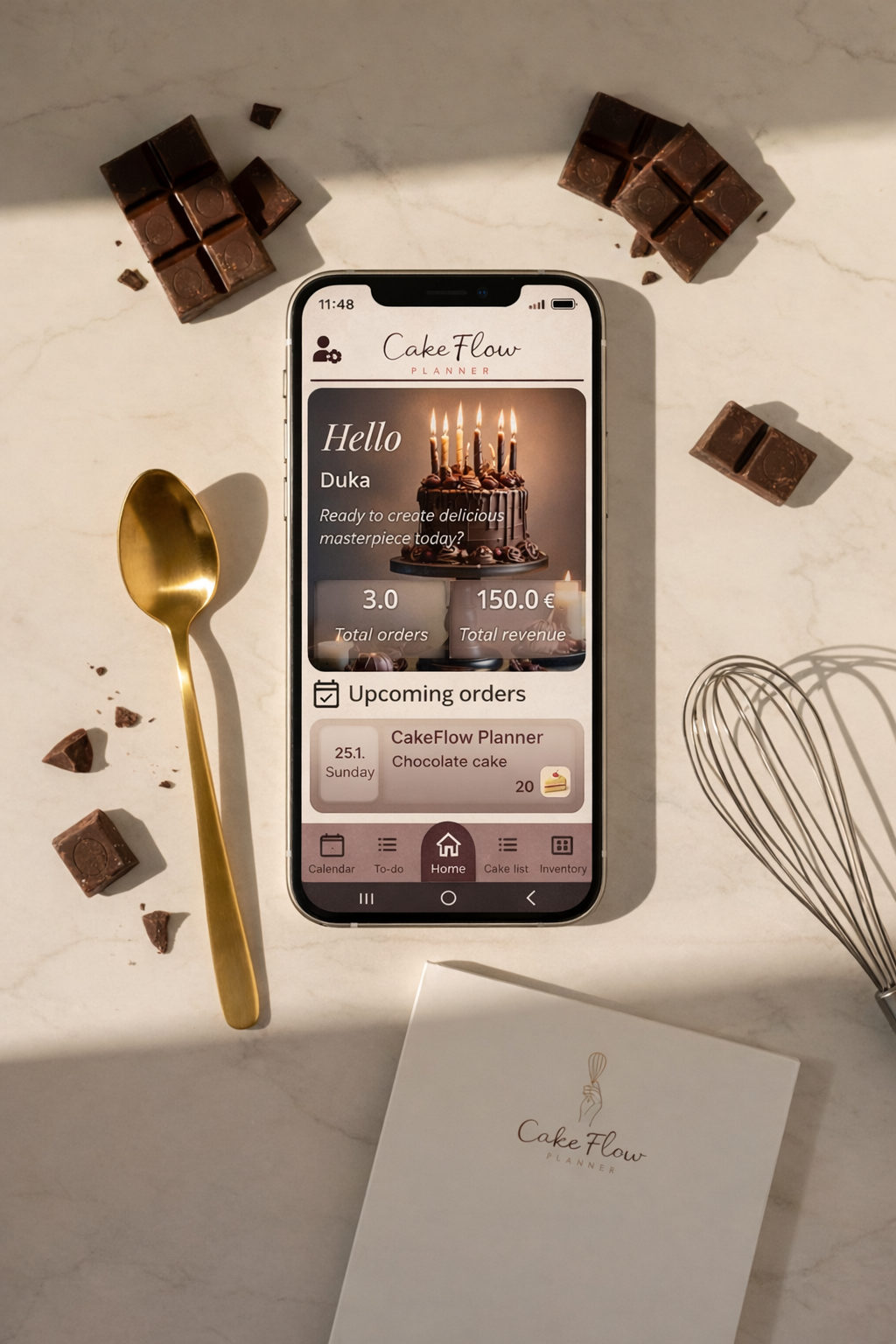 Smartphone displaying CakeFlow planner app on a marble countertop, surrounded by broken chocolate pieces, a gold spoon, and a wire whisk, with a CakeFlow note pad below.