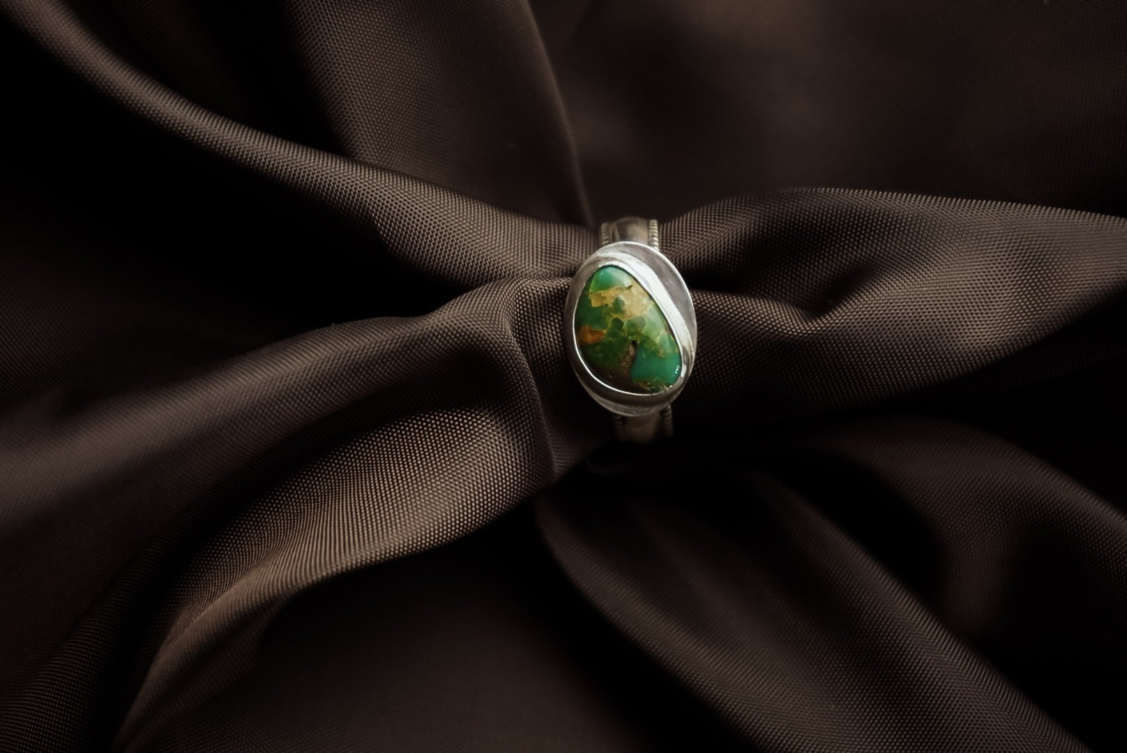 A silver ring with a large, oval green and yellow gemstone, resting on dark brown fabric.