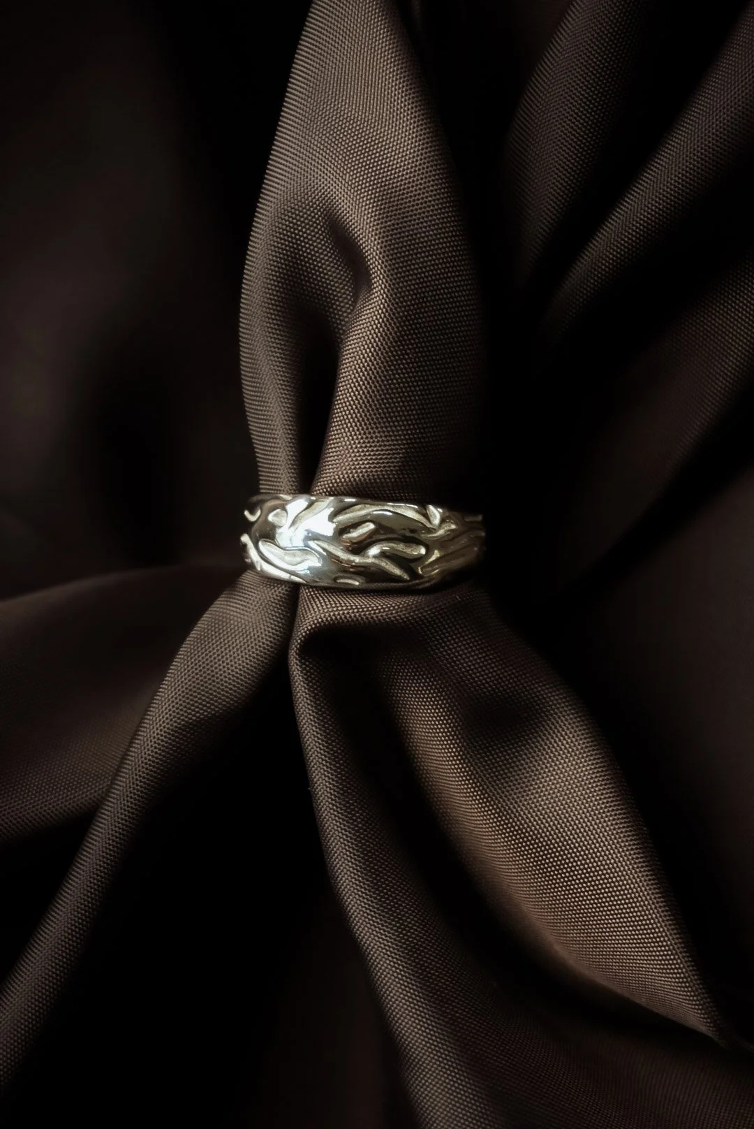Close-up of a silver ring with a flame design, placed on dark brown satin fabric.