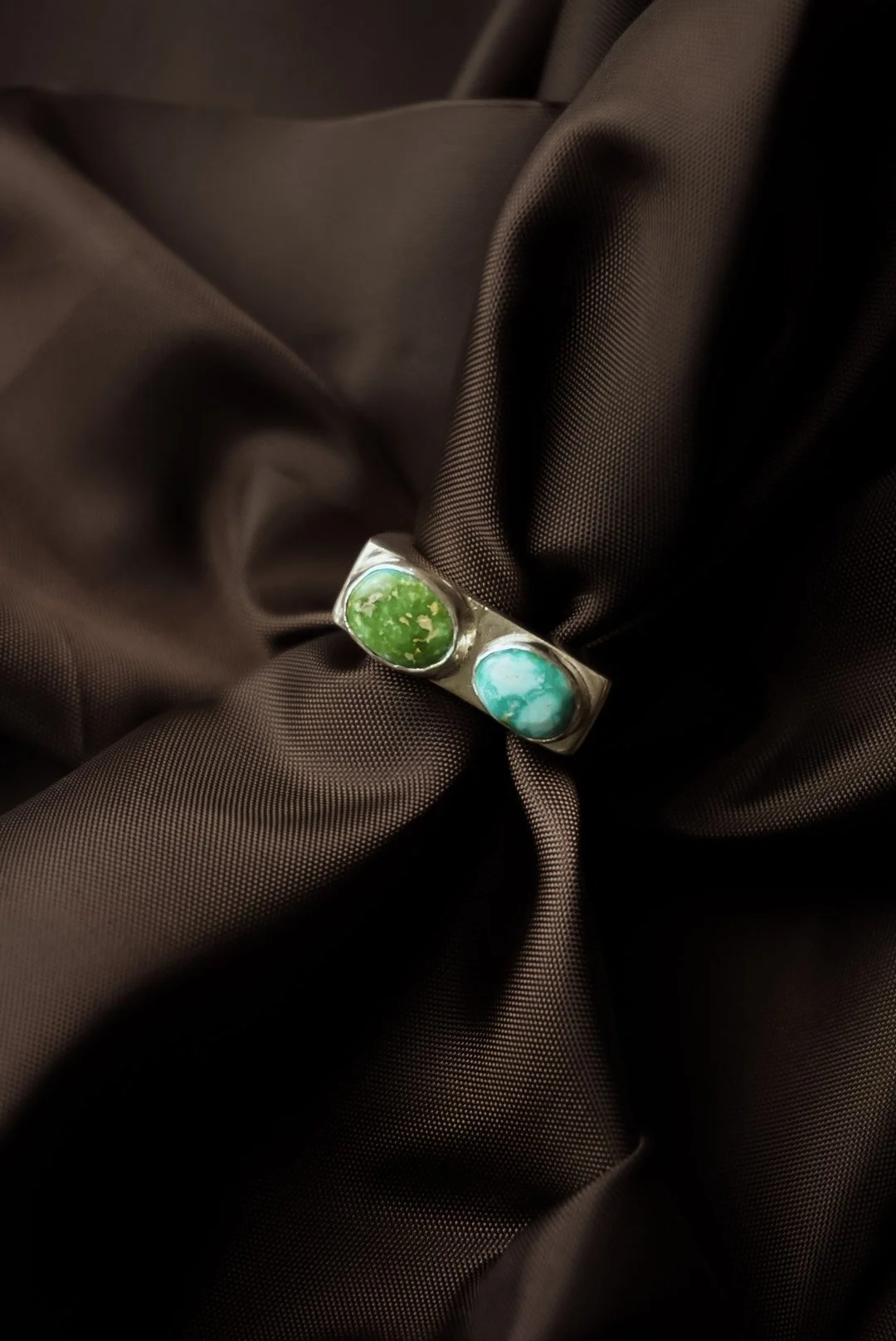 A silver ring with two oval turquoise stones, one green and one blue, resting on dark brown fabric.