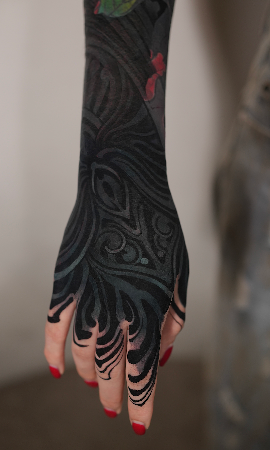 A person's hand with red painted nails, showing a black sleeve tattoo with red accents and intricate swirling patterns.