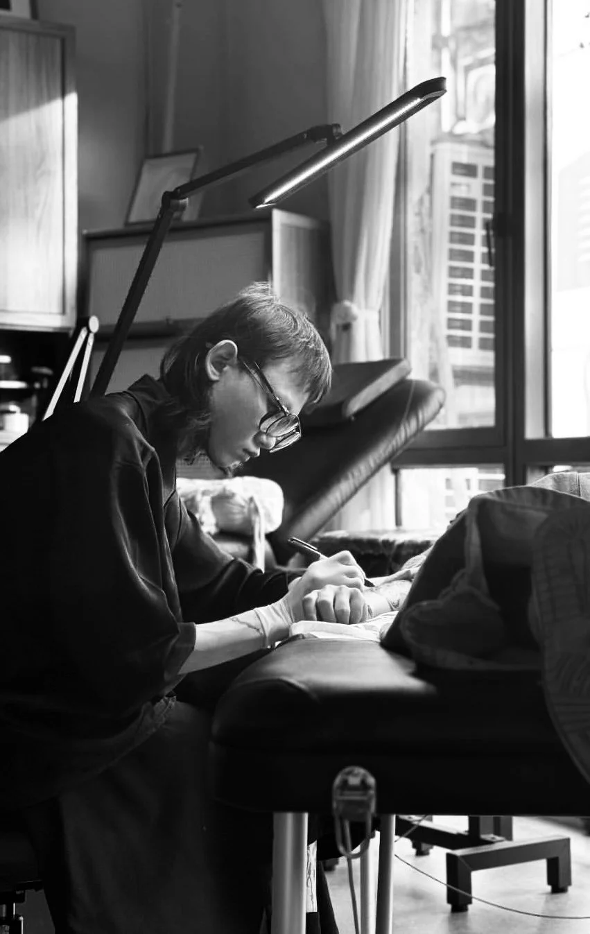 A person with glasses writing on a piece of paper at a desk in a room with sunlight coming through a window.