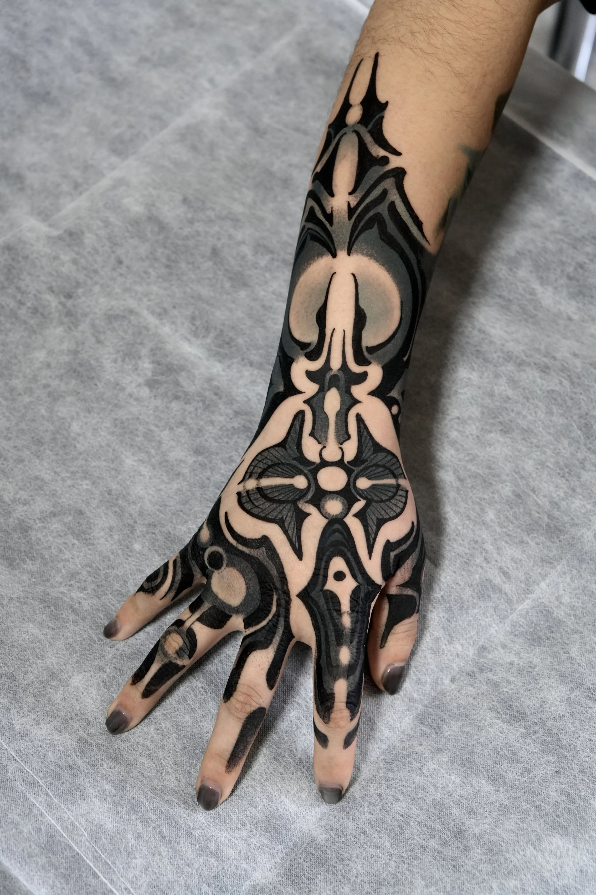 Close-up of a person’s hand and forearm with intricate black and beige tattoo artwork covering the skin.