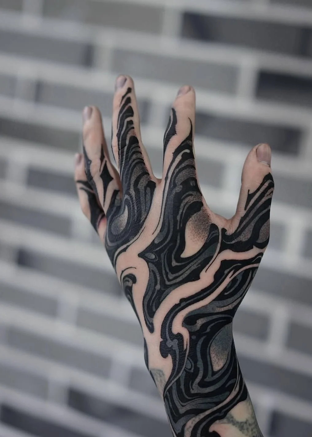 A hand with black and pink marbled tattoos against a blurred gray metal background.