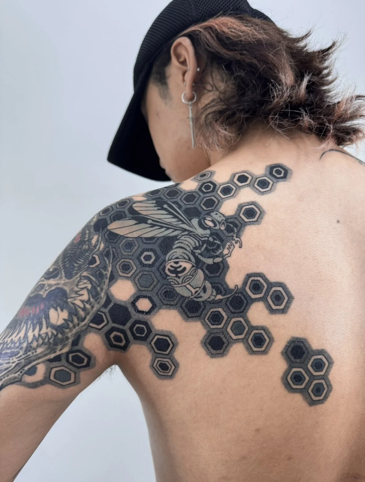 A person with short hair, wearing a black cap, has a large tattoo of a bee on their shoulder and upper back, surrounded by a honeycomb pattern.