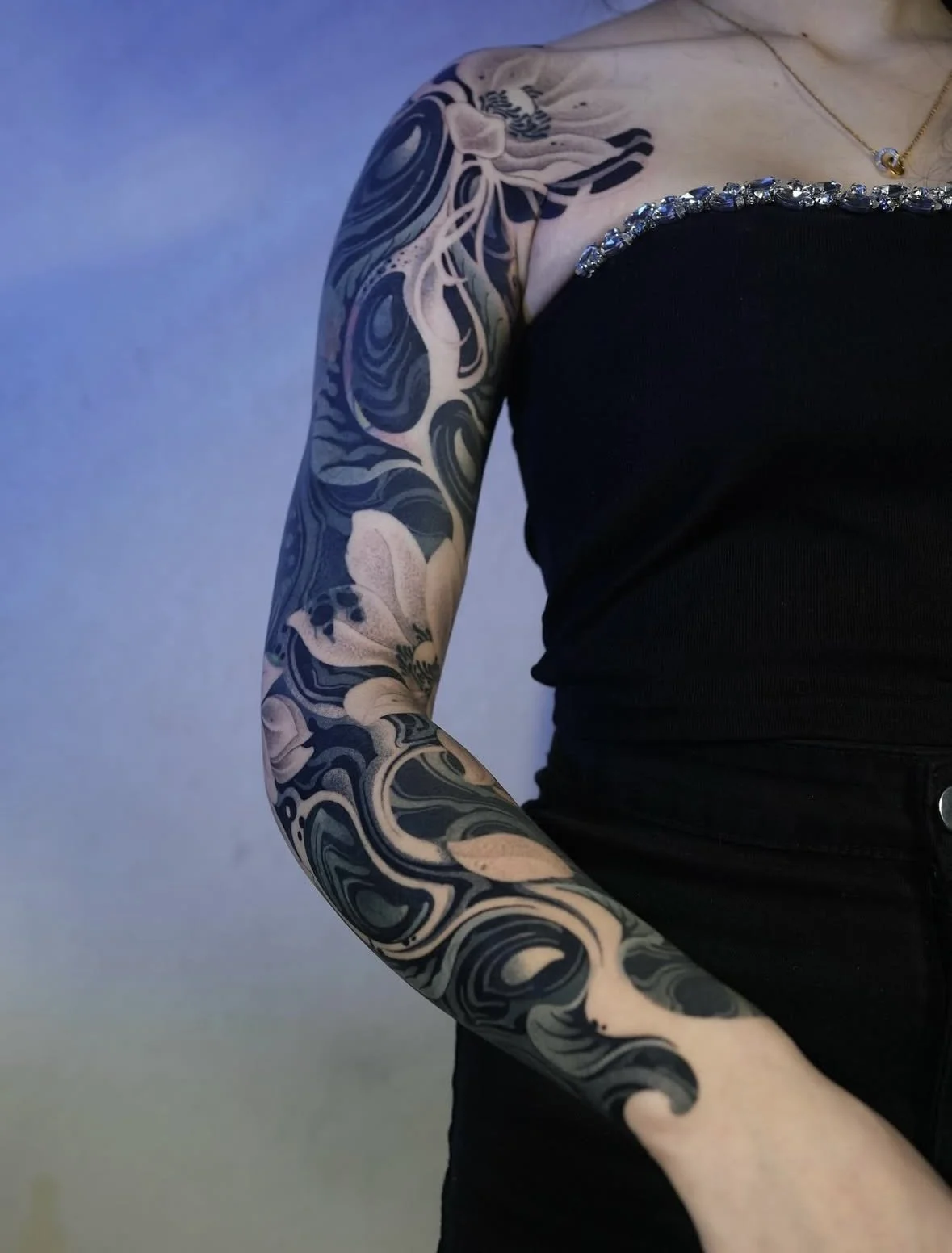 Close-up of a person's intricately designed black and gray sleeve tattoo on their arm, wearing a black sleeveless top with rhinestones along the shoulder, and a delicate gold necklace, against a plain blue background.