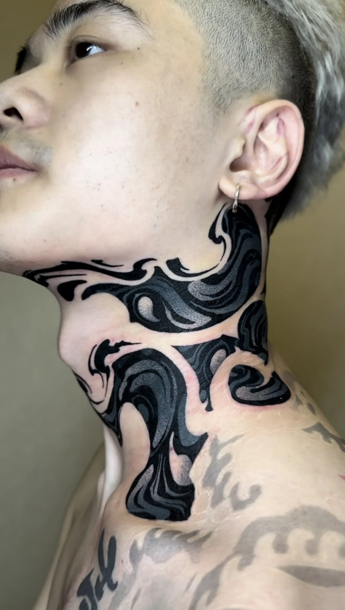 Close-up of a person's side profile showing short, styled hair, an earring, and a large black and pink tribal tattoo covering the neck and part of the shoulder.