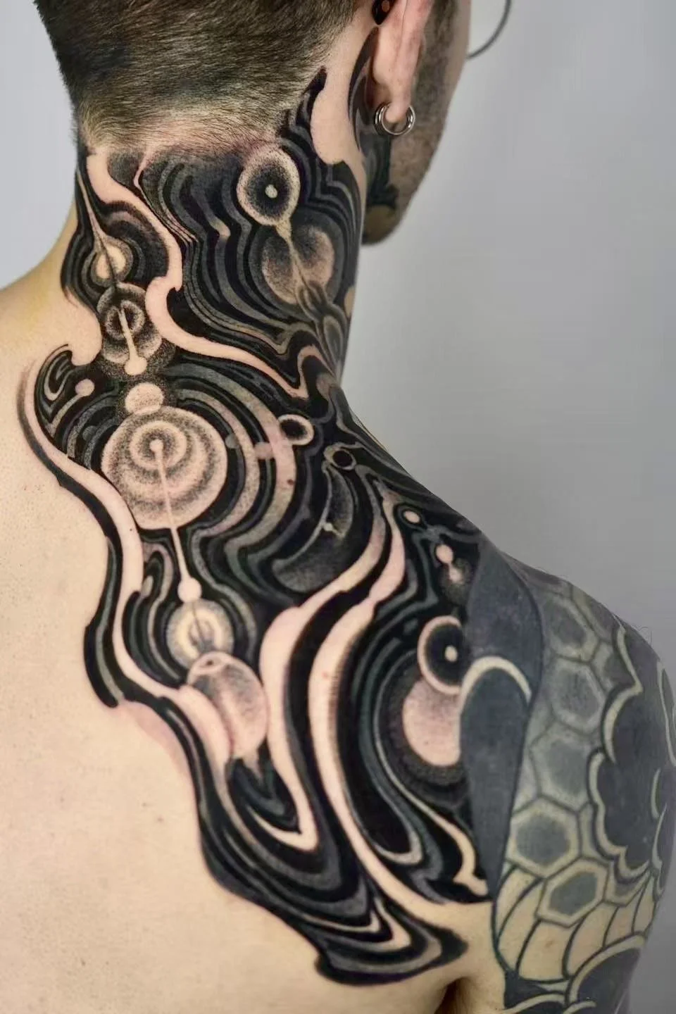 Close-up of a person's upper back and neck showing a black and pink geometric and abstract tattoo design, including circular, wavy, and honeycomb motifs.