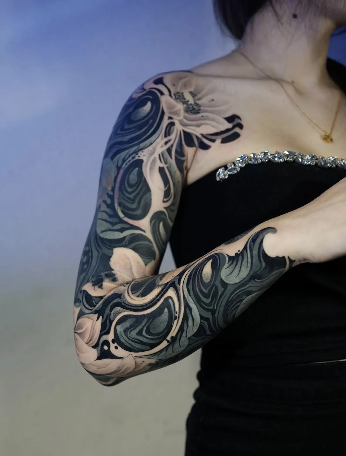 Close-up of a woman's arm and shoulder with a black and white tattoo sleeve featuring swirling patterns and abstract designs, wearing a black top with decorative rhinestones and a gold necklace.
