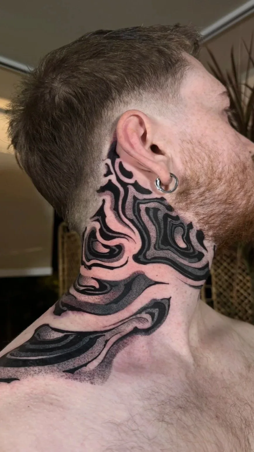 A shirtless man with short brown hair and a large hoop earring in his left ear, showcasing an intricate black and pink tattoo design that covers his neck and part of his shoulder.