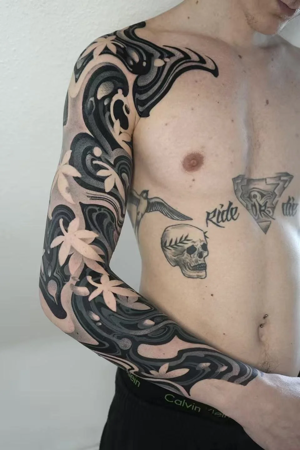 A person with tattoos, including a sleeve of black and beige floral designs on the arm, a skull with a leaf branch on the chest, a swallow bird, a Superman logo, and the words "ride" and "the" on the torso.